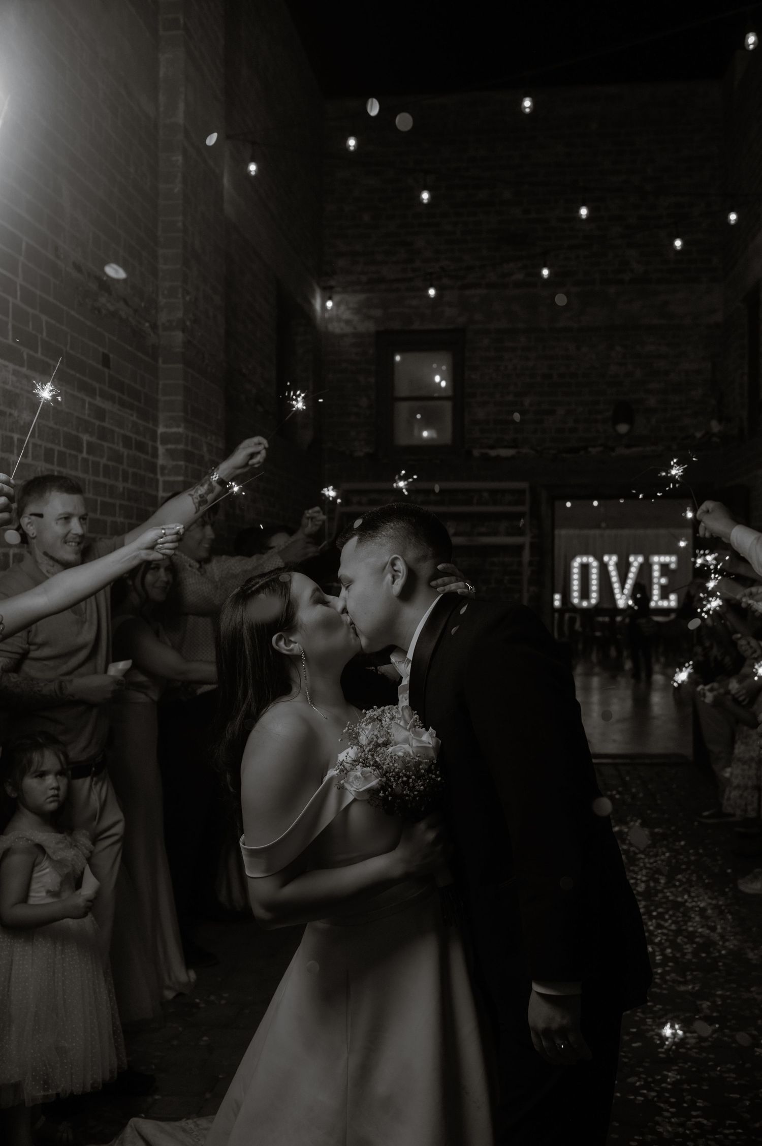 Black-and-white photo of bride and groom kissing on dance floor, with “LOVE” marquee glowing in the background.