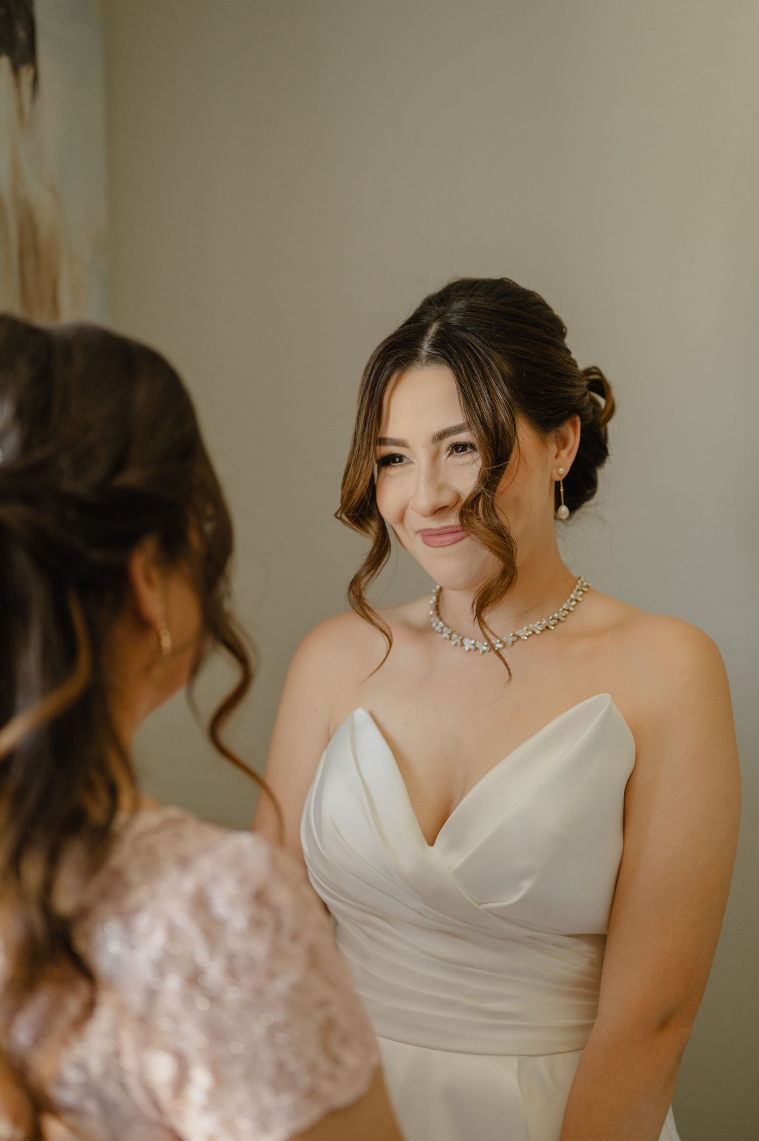 Bride smiles while speaking with bridesmaid in bridal suite.