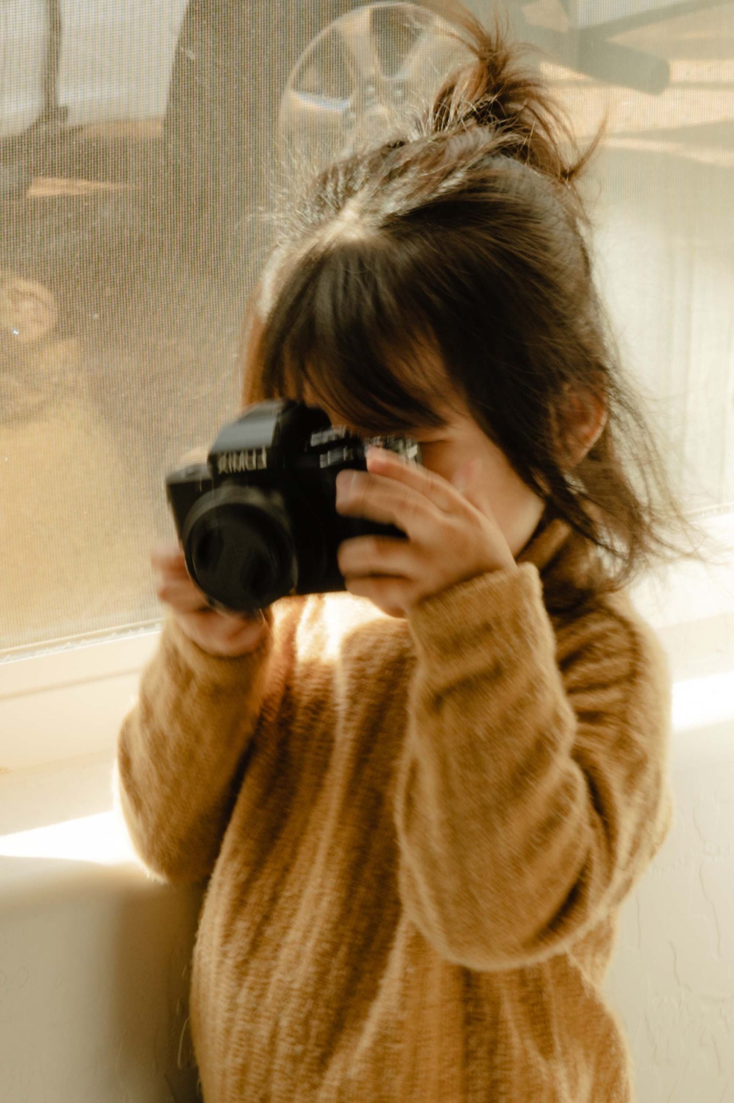 Young child in tan sweater holding a DSLR camera up to her face near a window.