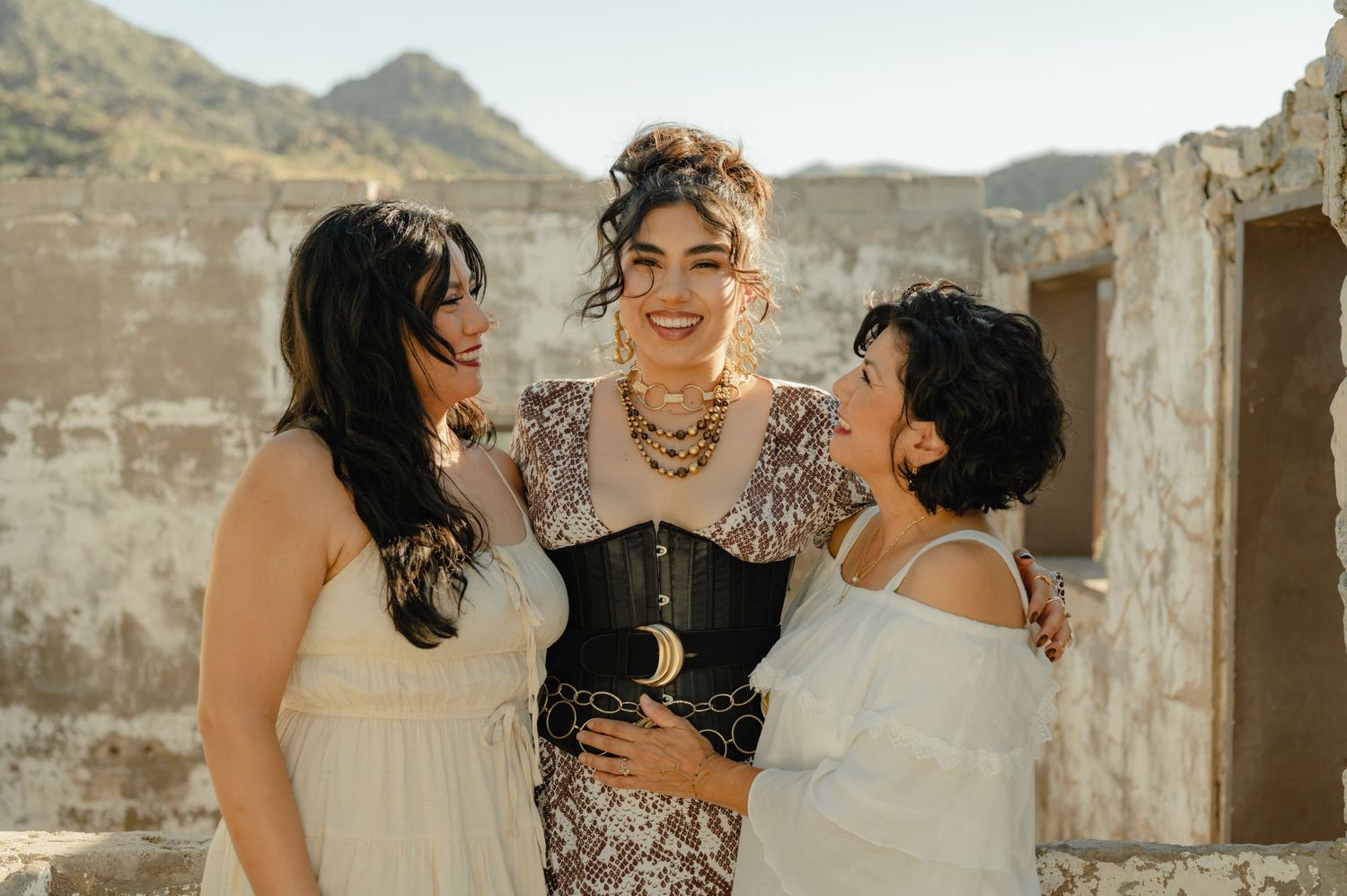 Three women laugh and chat together outdoors at a rustic desert location.