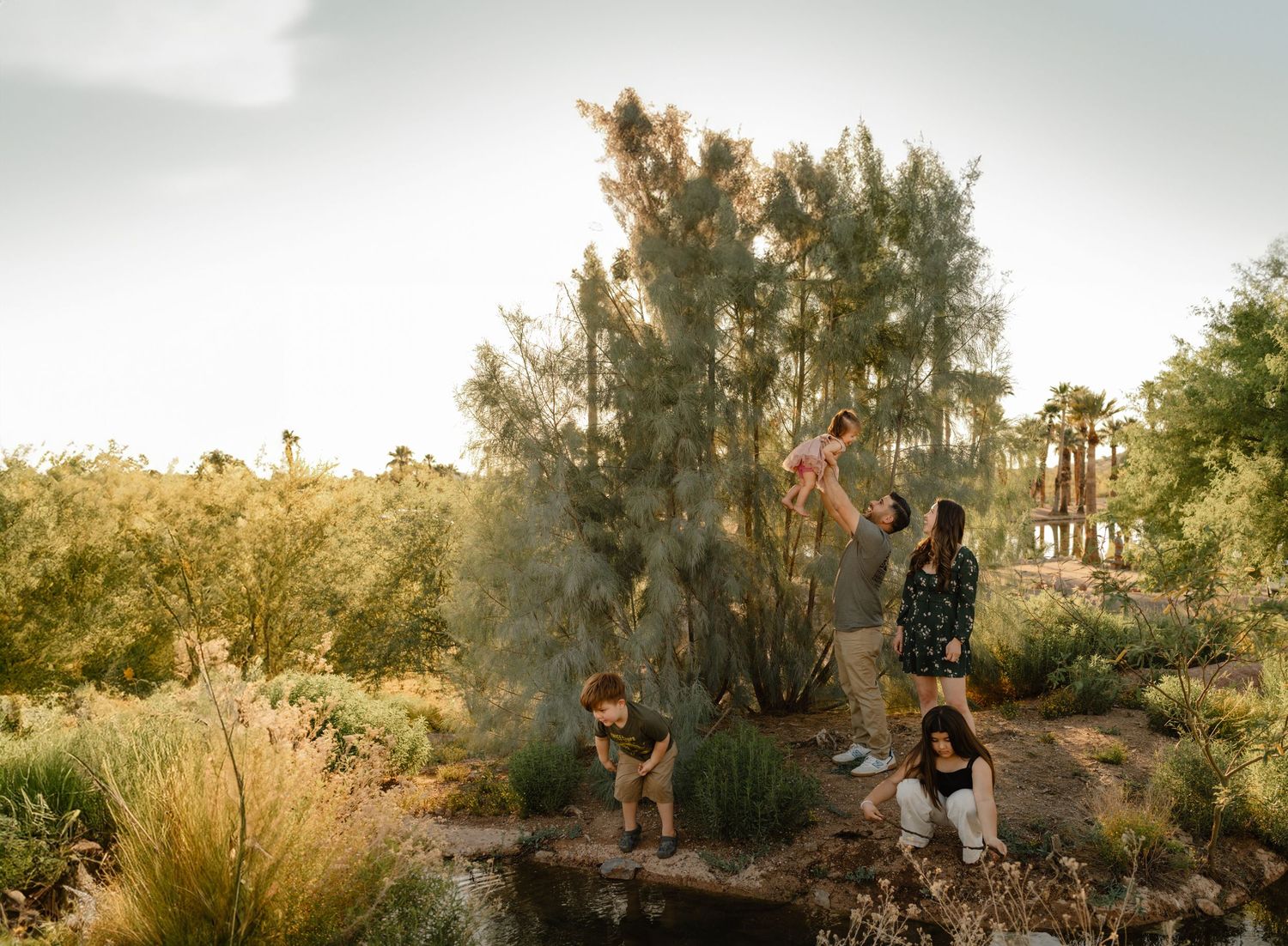 A family plays together near desert trees at dusk in Papago Park, Phoenix, AZ, a parent lifting a child in golden light.