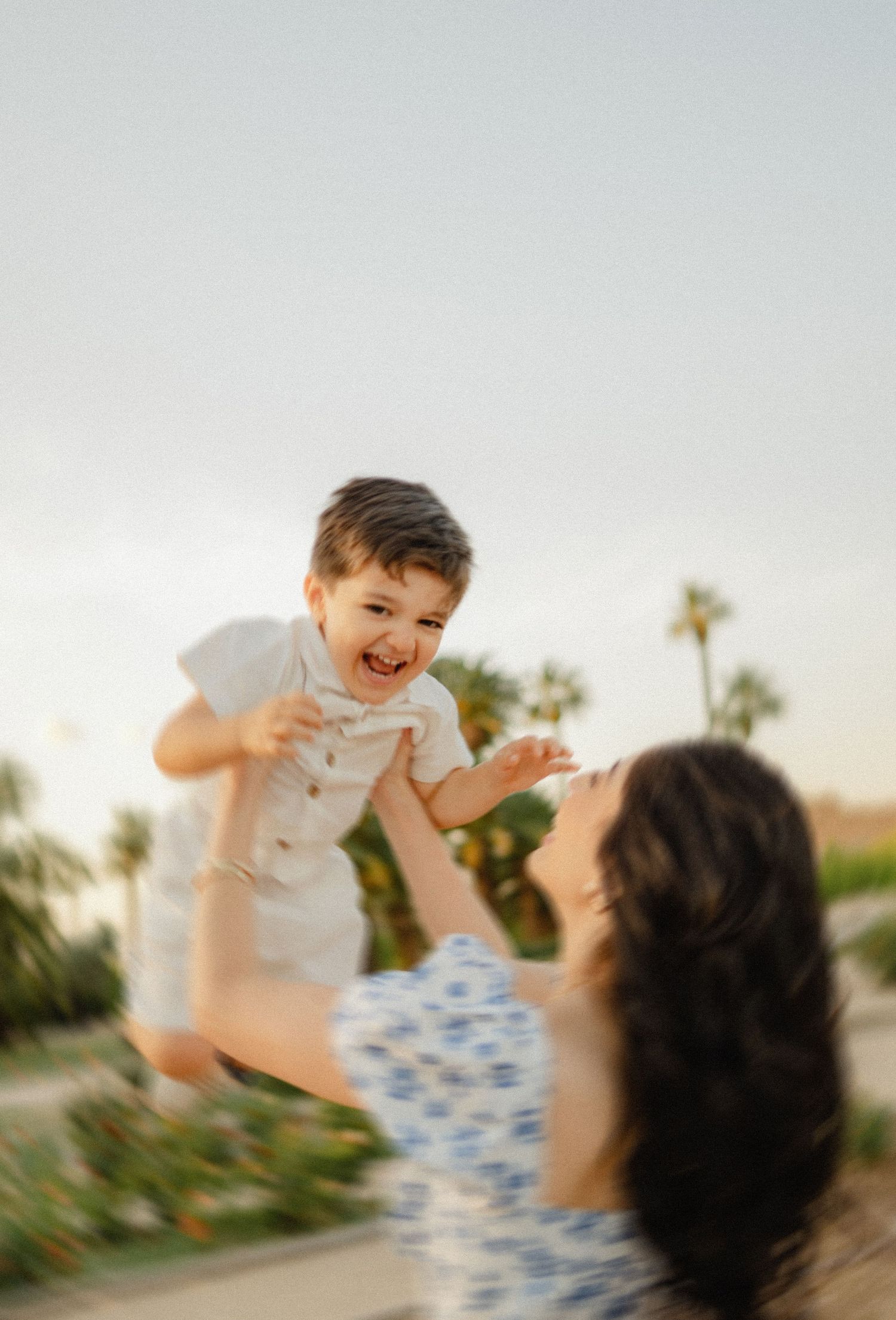 A joyful toddler is lifted into the air by his mother at Sahuaro Ranch Park in Glendale, AZ, palm trees framing the sky.