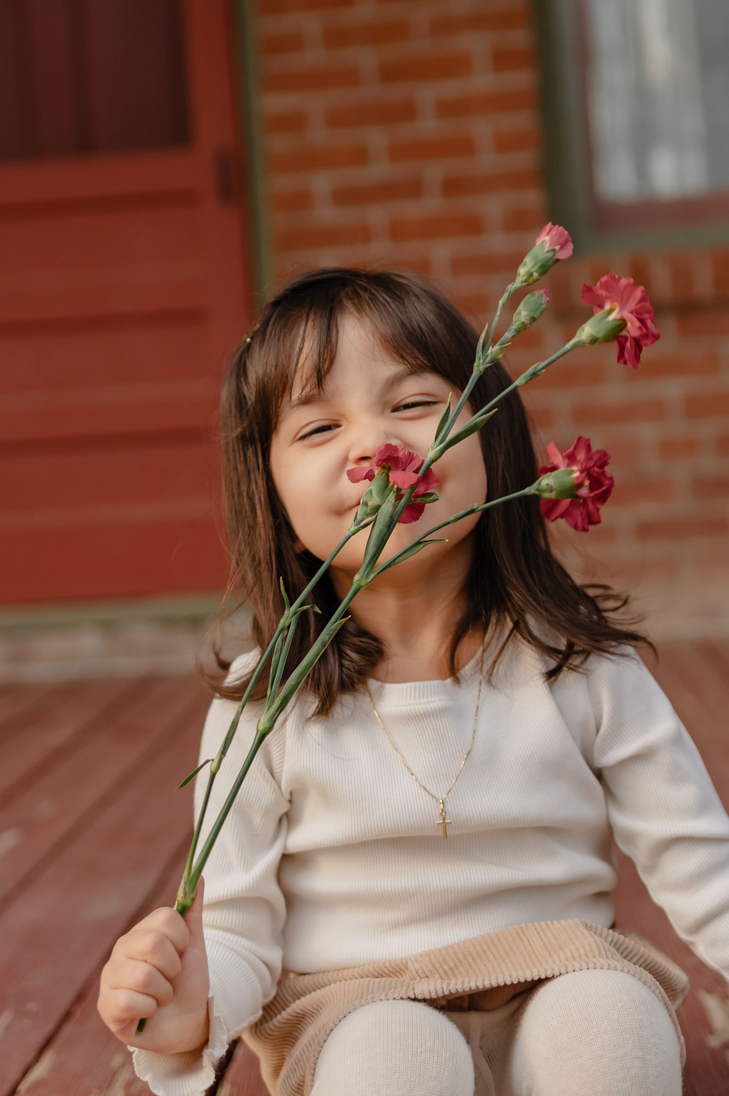 A young girl in white makes a kissing face while holding pink carnation flowers.