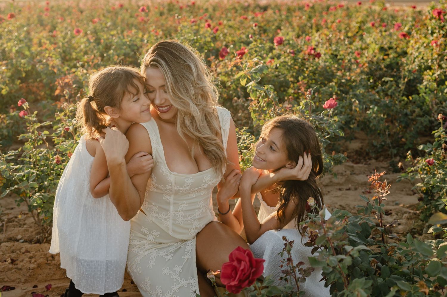 Blonde mother with two daughters in white dresses in blooming rose field.