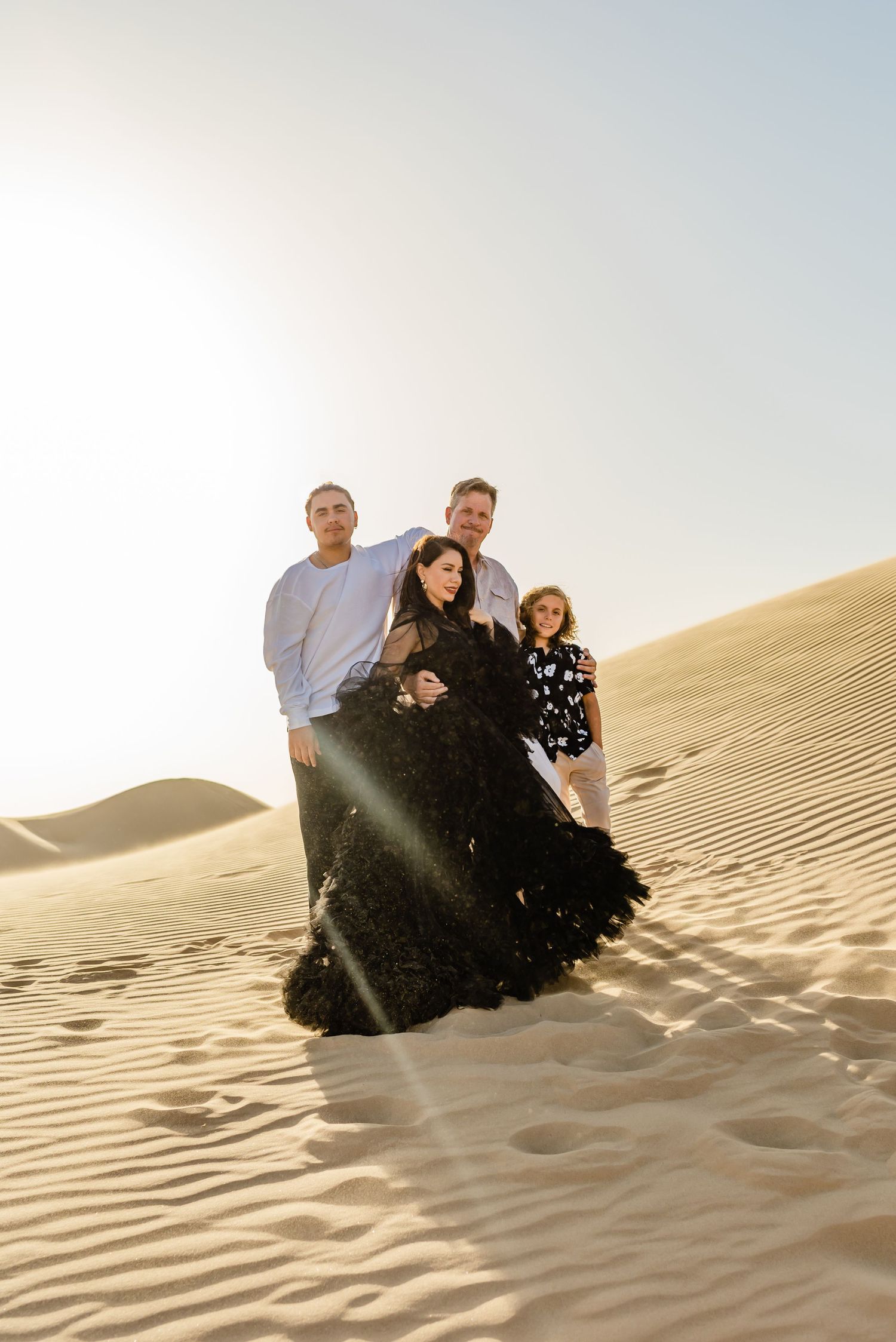 Family of four posing on golden sand dunes in the desert, woman in elegant black gown at golden hour.