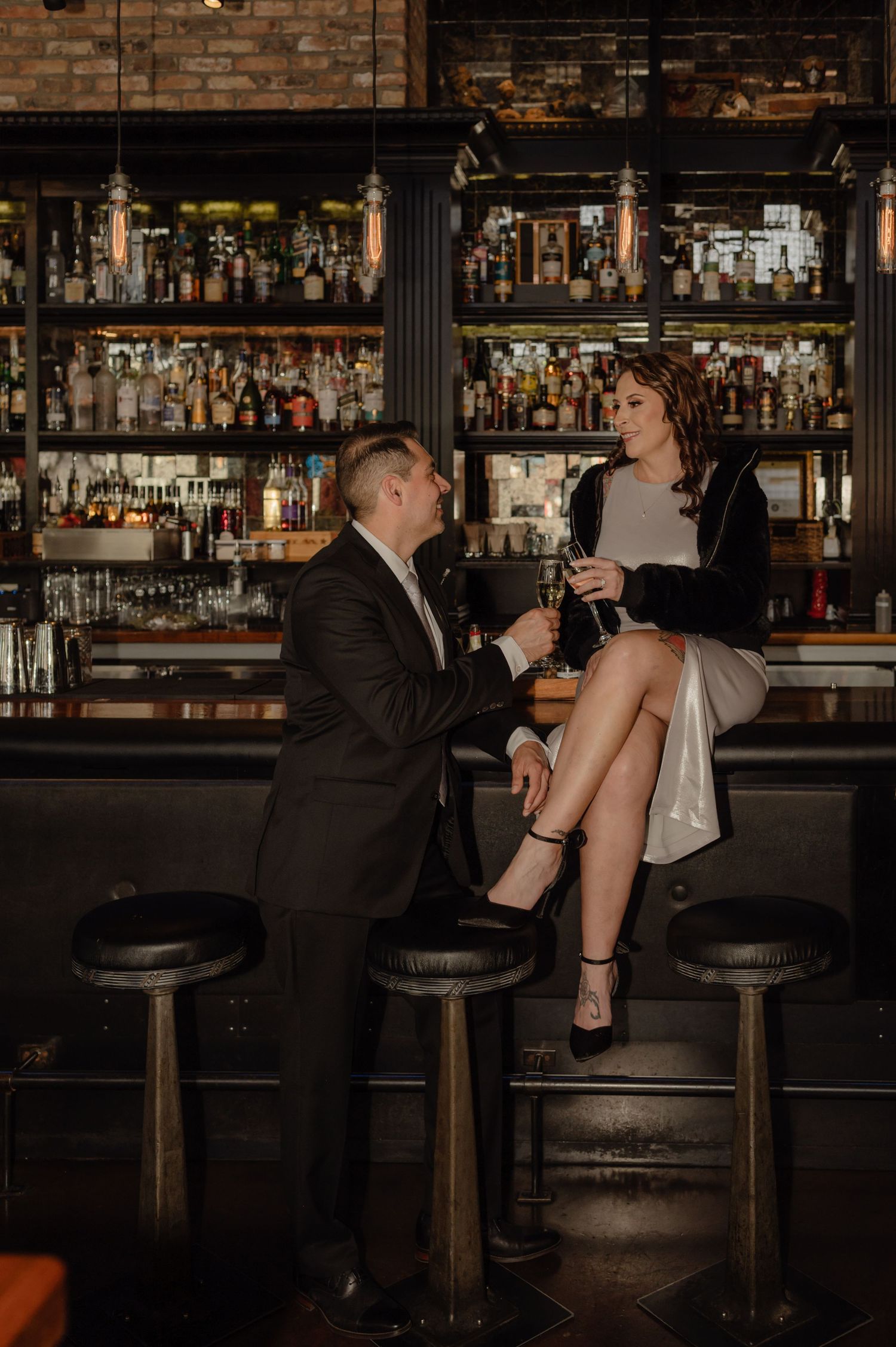Couple sharing a romantic toast at a dimly lit bar, man in suit kneeling beside woman seated on barstool.