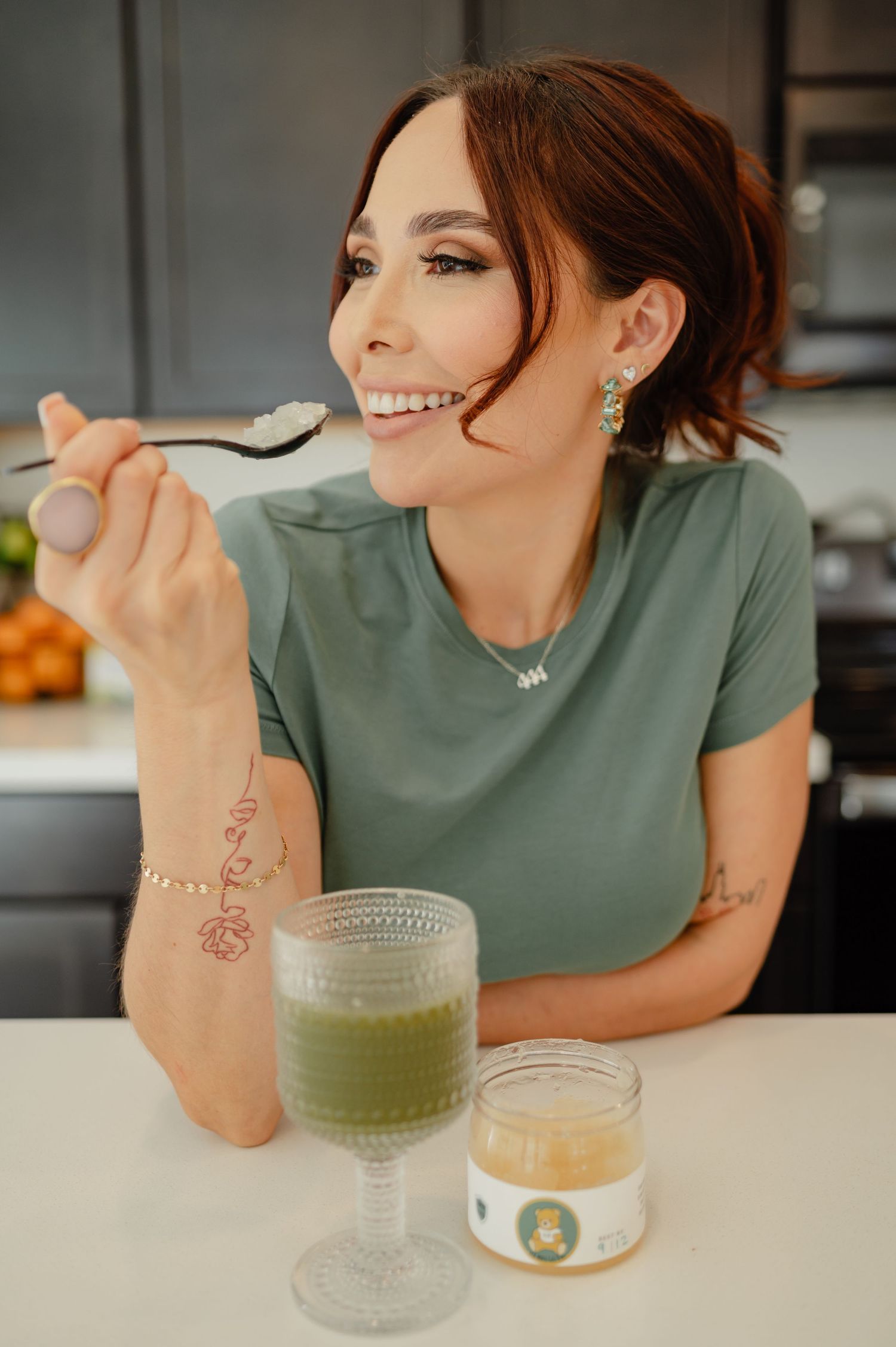 Smiling woman in green shirt enjoying a spoonful of supplement near a green drink.