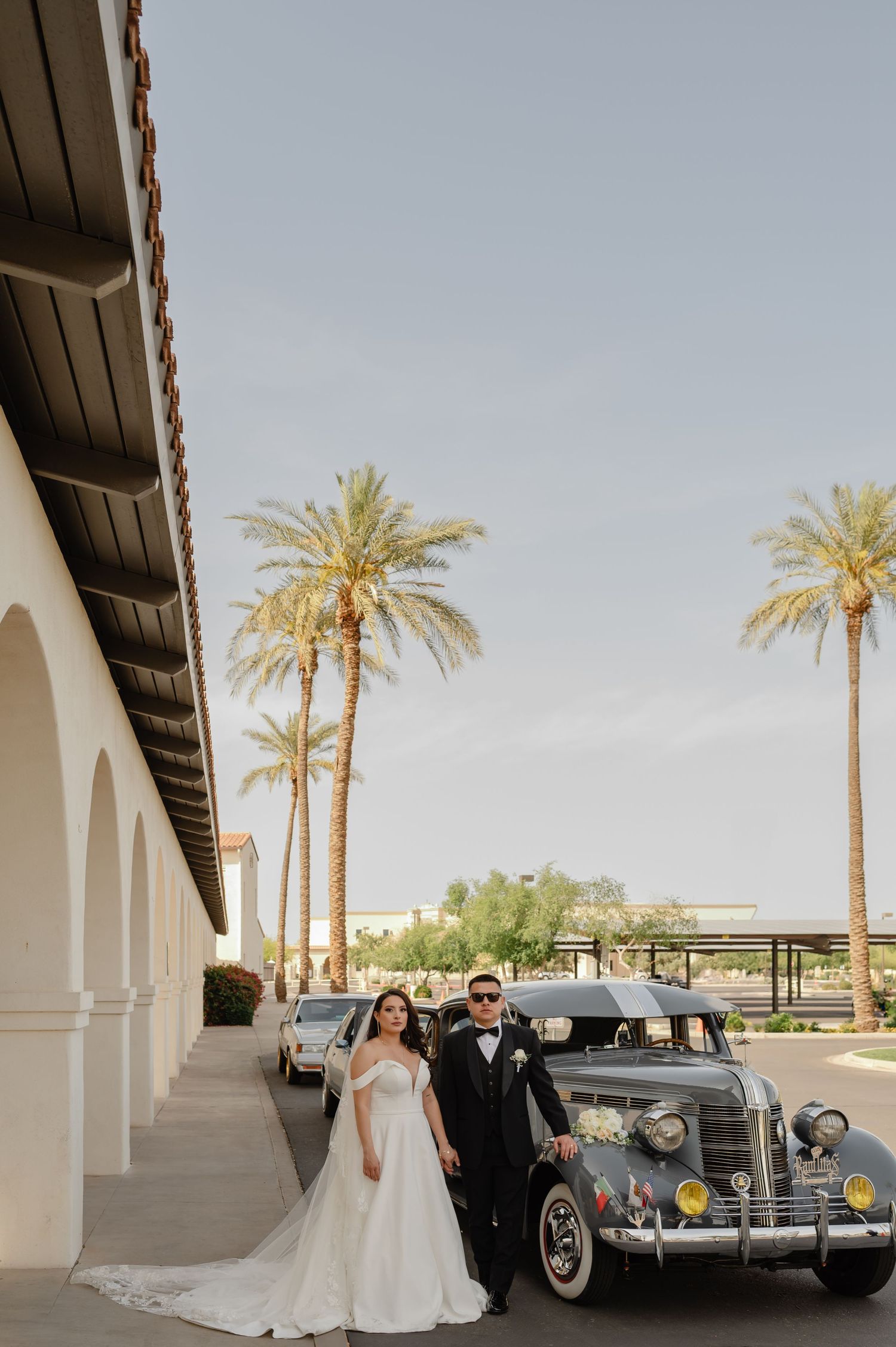 Bride and groom pose in front of vintage car outside Tuscan-style venue with palm trees and arches.