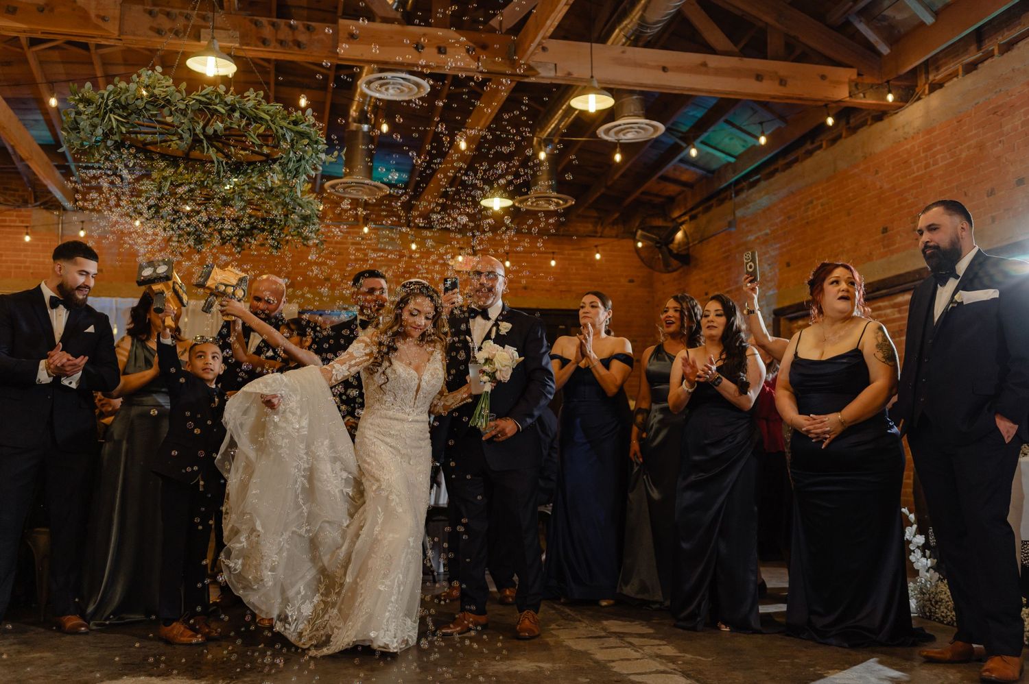 Wedding recessional with joyful confetti toss, bride and groom smiling as guests cheer, captured in warm indoor light.