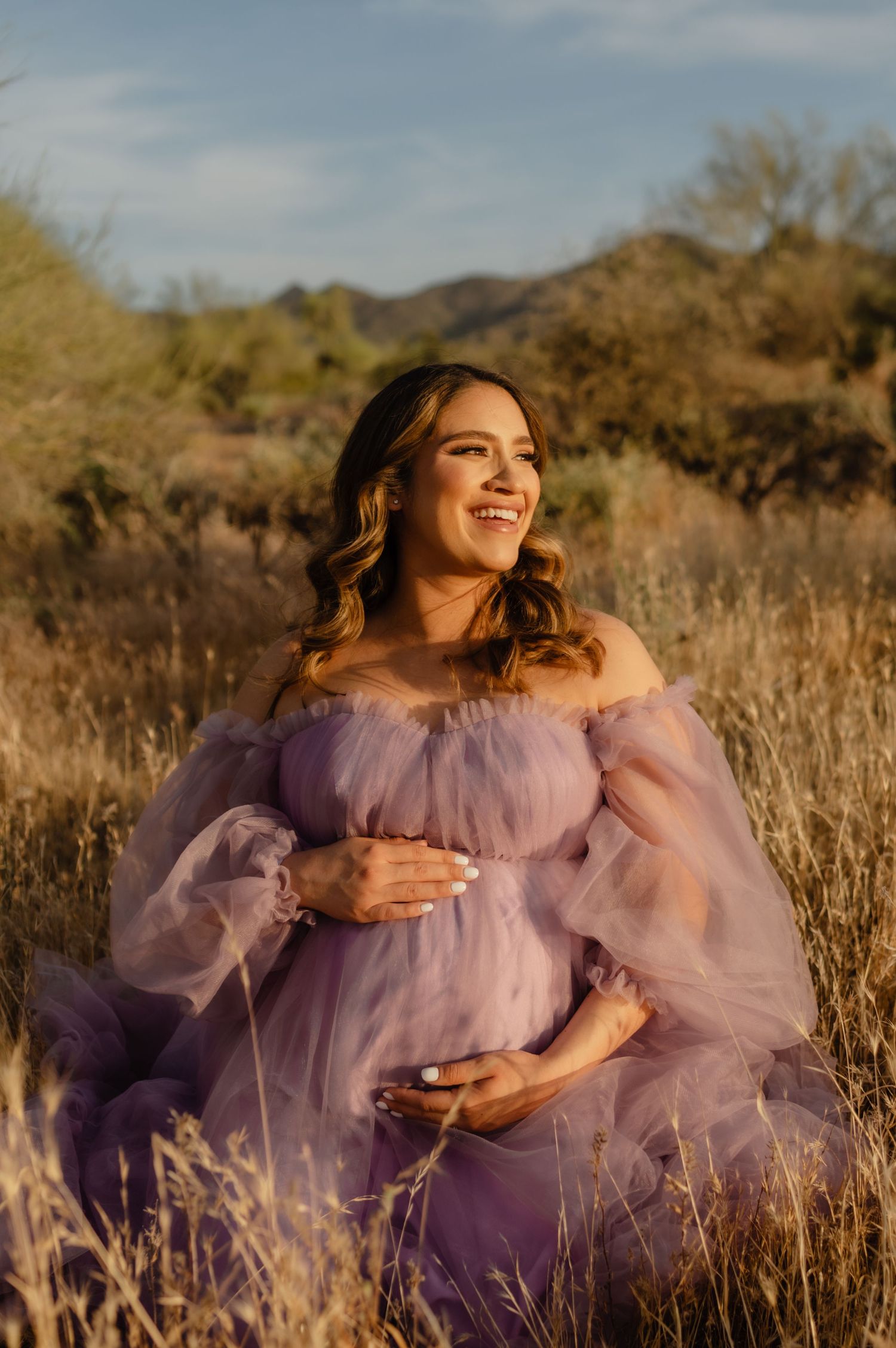 pregnant woman smiles in a purple tulle gown surrounded by golden grasses during a phoenix desert session