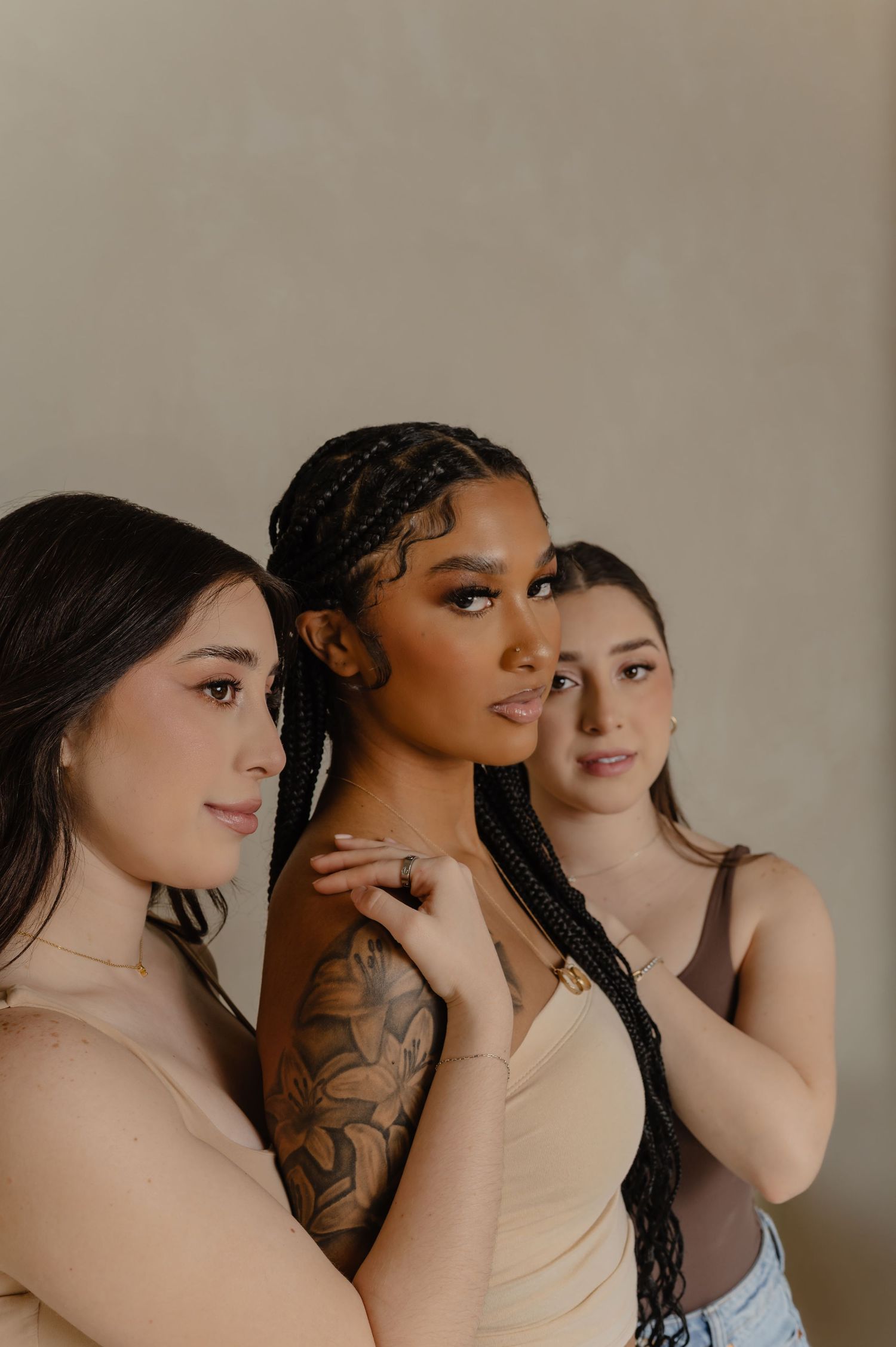 Three women pose together in elegant makeup, one with braided hair and a delicate tattoo, dressed in neutral tones.