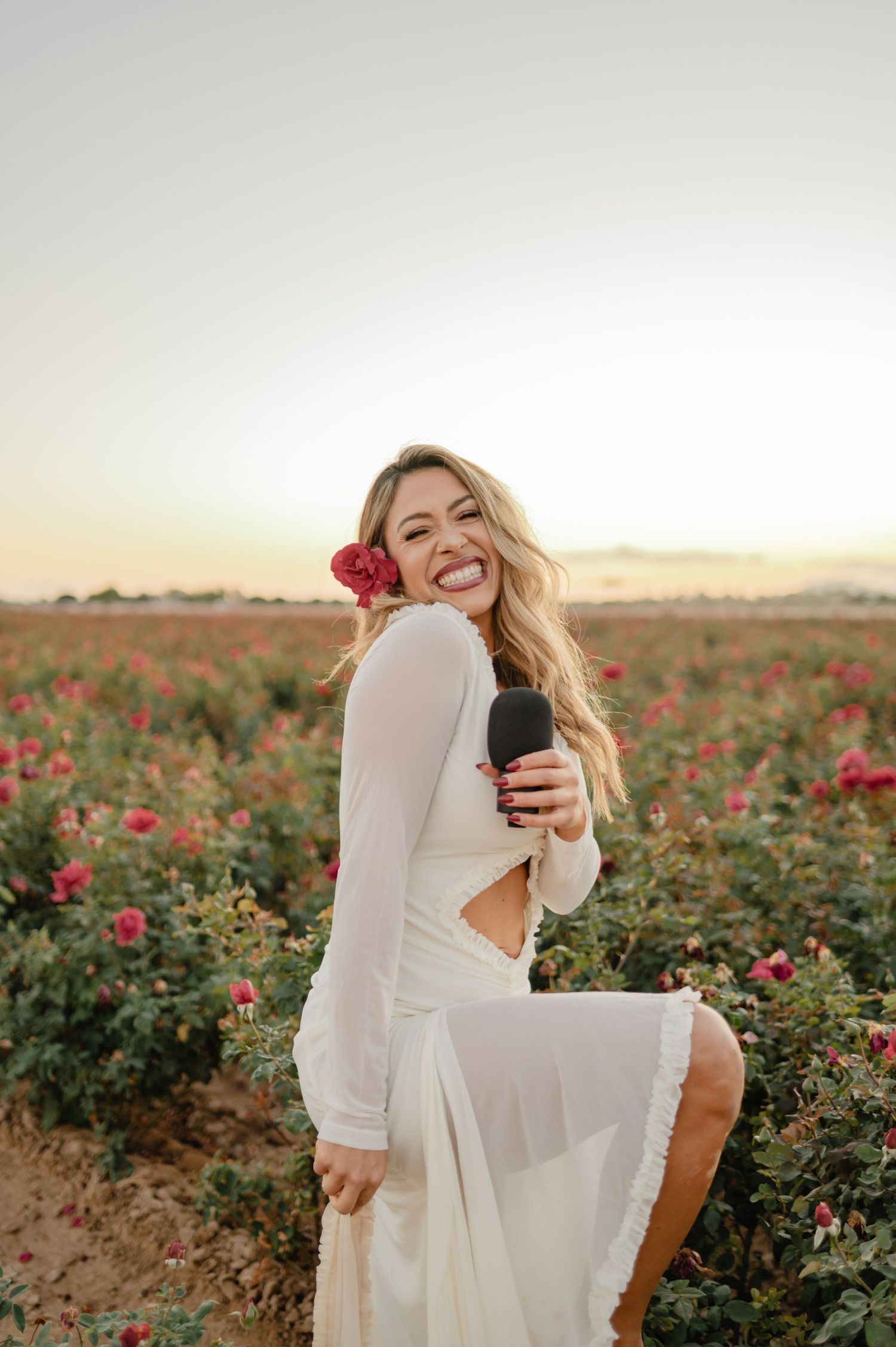 Rosy Liddi, founder of Calma Wellness Lounge, smiles with a red flower tucked in her hair amid blooming roses at sunset.