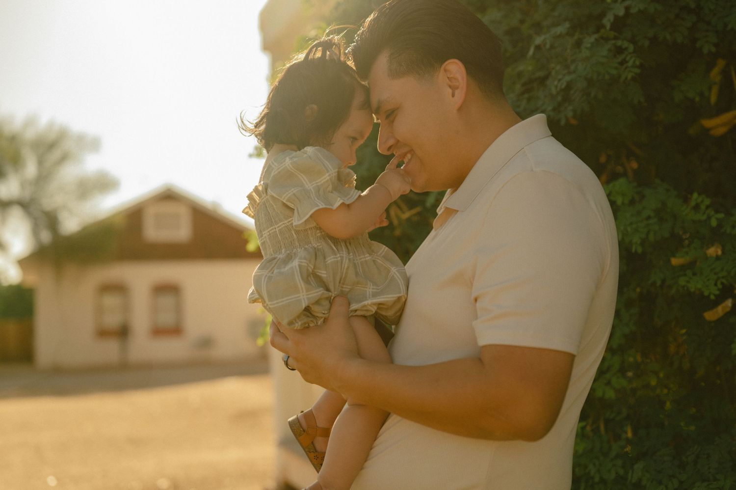 Father tenderly holding baby daughter outdoors in warm golden sunset light near rustic building.