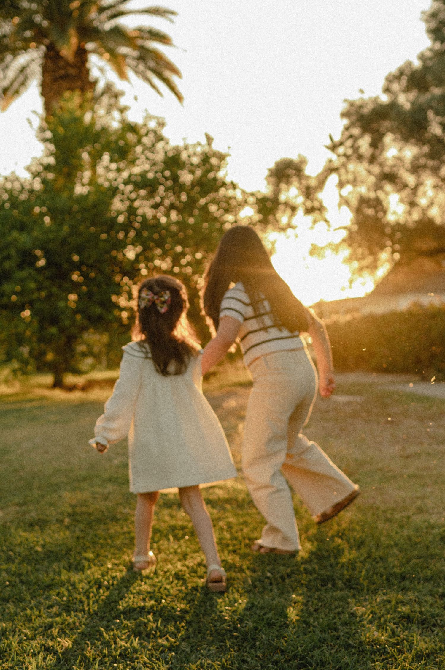 A mother and young daughter hold hands dancing on green grass beneath a palm tree at sunset.