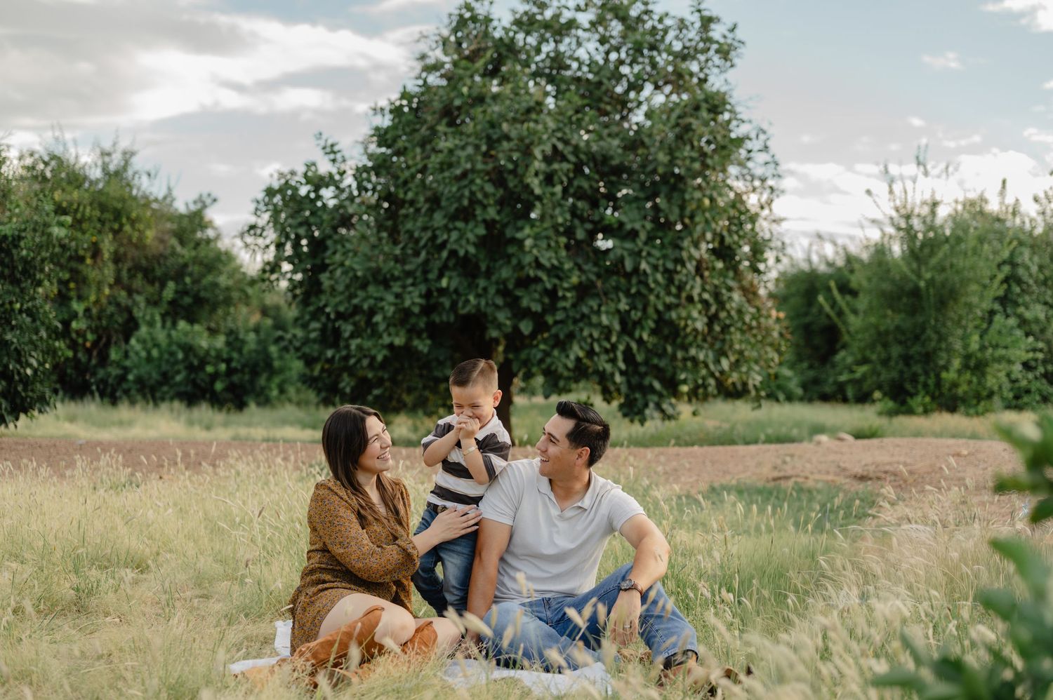 Happy family of three enjoying a picnic in a lush green meadow with trees in the background.