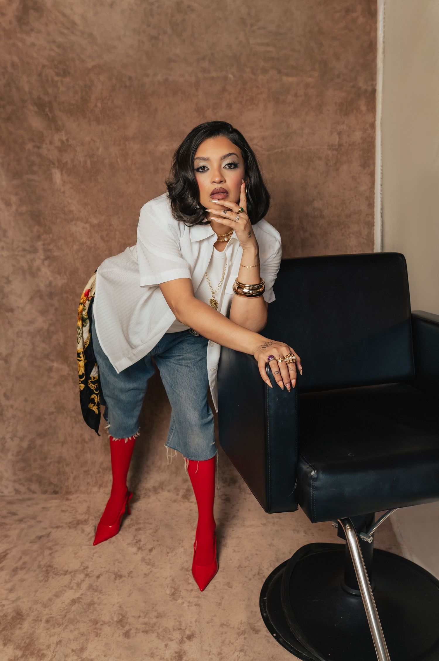 Seated portrait of a woman adjusting her white blouse, red heels resting on the floor — taken at The Hive Salon.