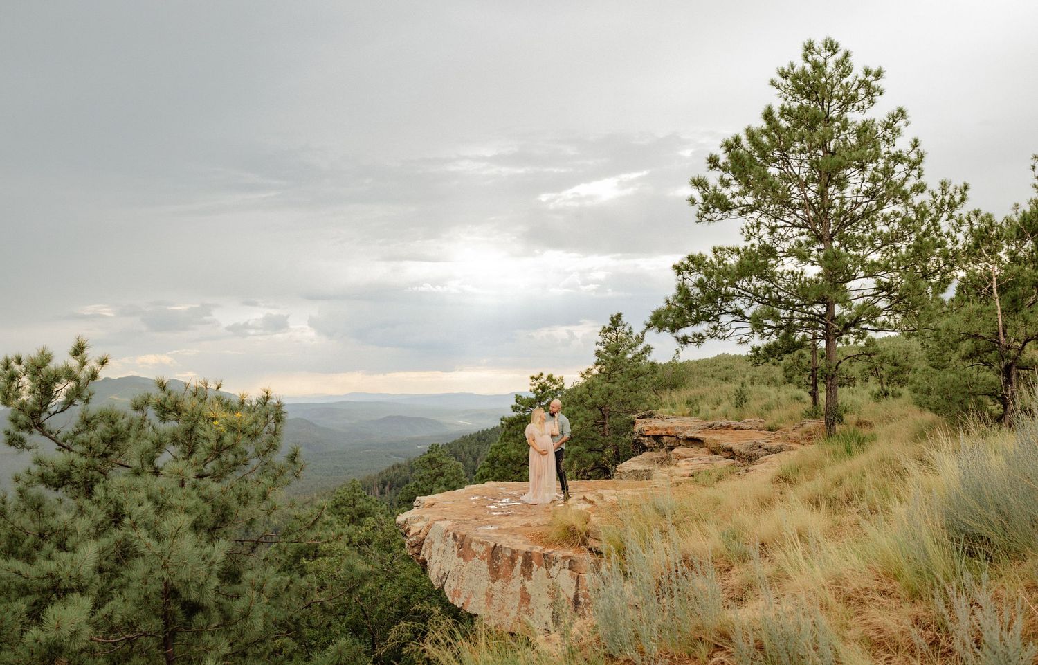 A bride stands on a rocky cliff edge surrounded by pine trees with a vast mountain valley view under a cloudy sky.