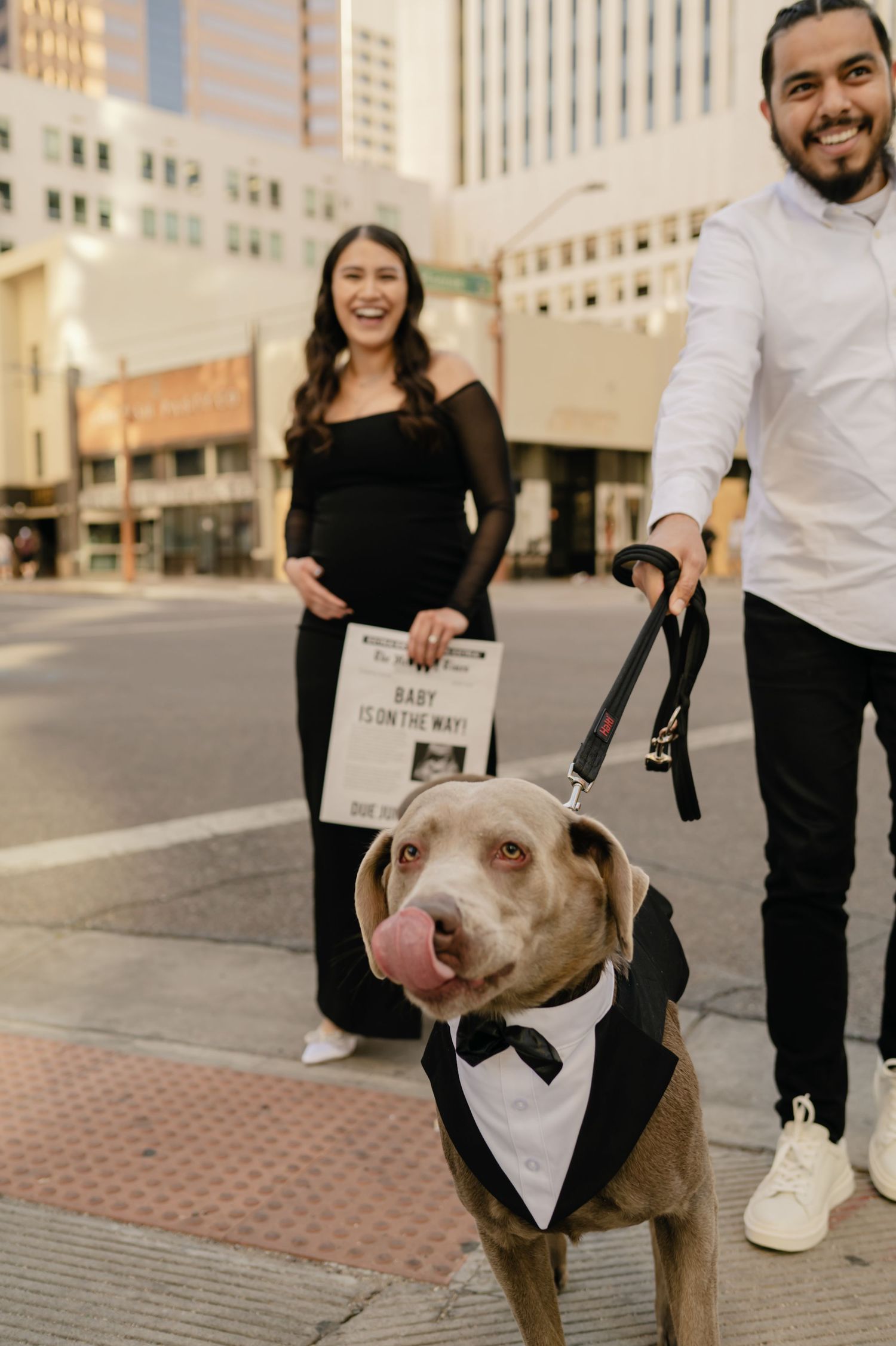 Couple in black and white outfits holds baby announcement sign with dog in tuxedo on city street.