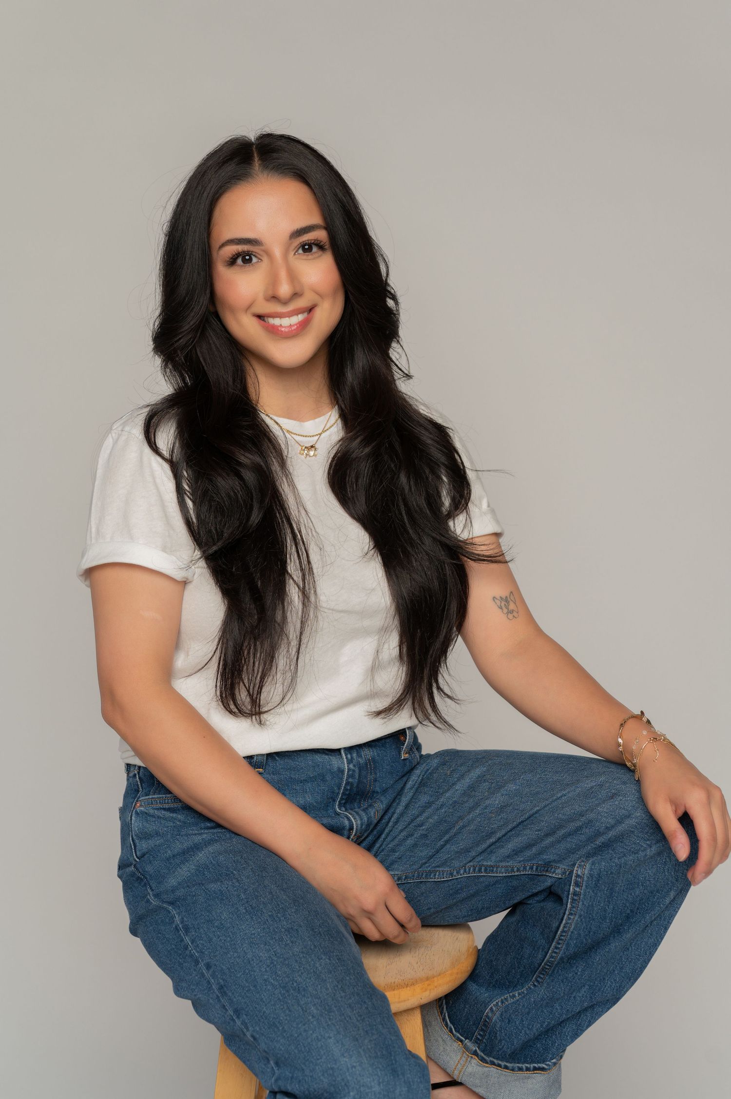 Young woman with long dark hair smiling, wearing white tee and jeans, seated on stool against gray background in studio in phoenix.