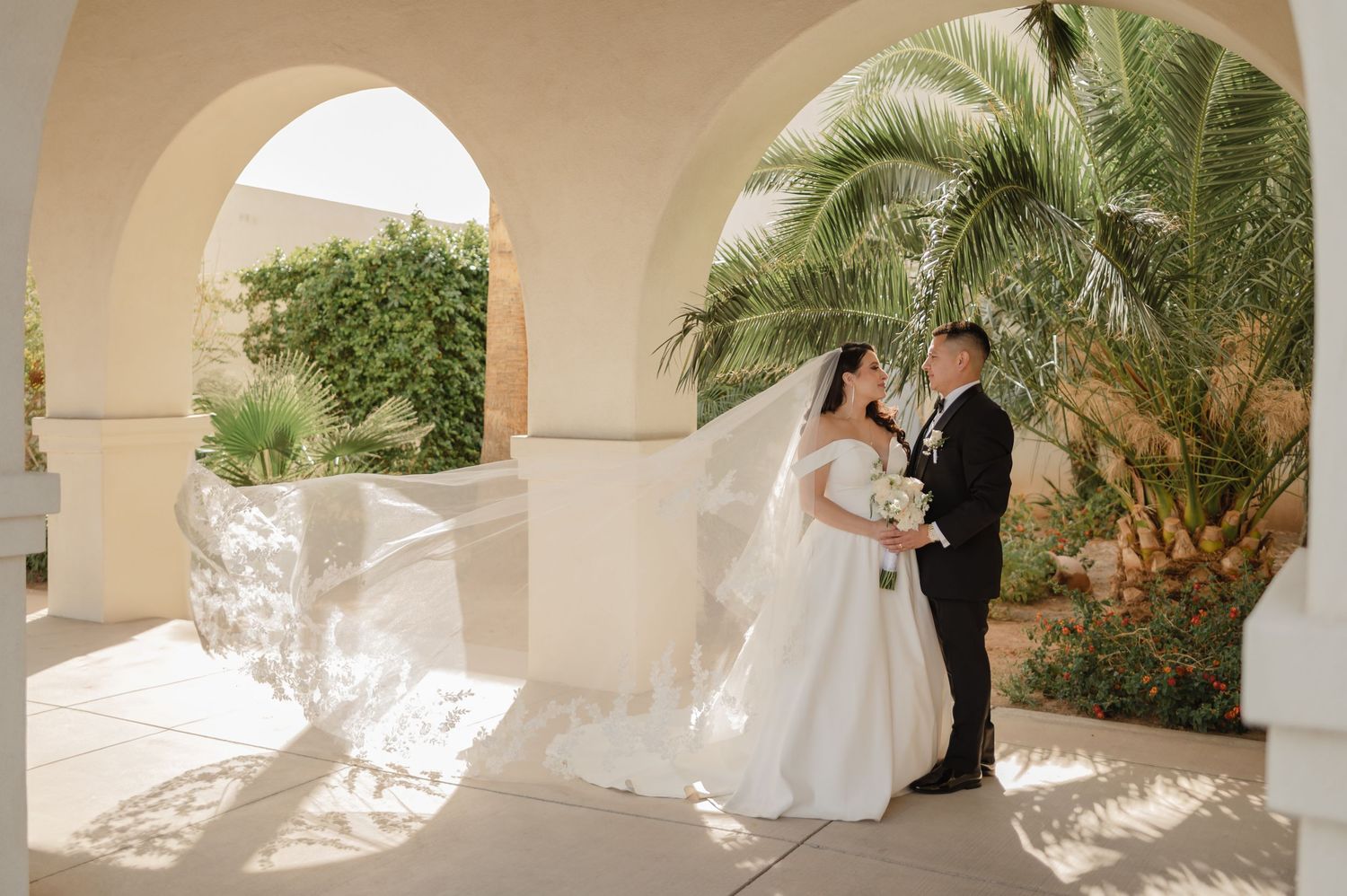Bride and groom share intimate moment under arched veranda with tropical greenery.
