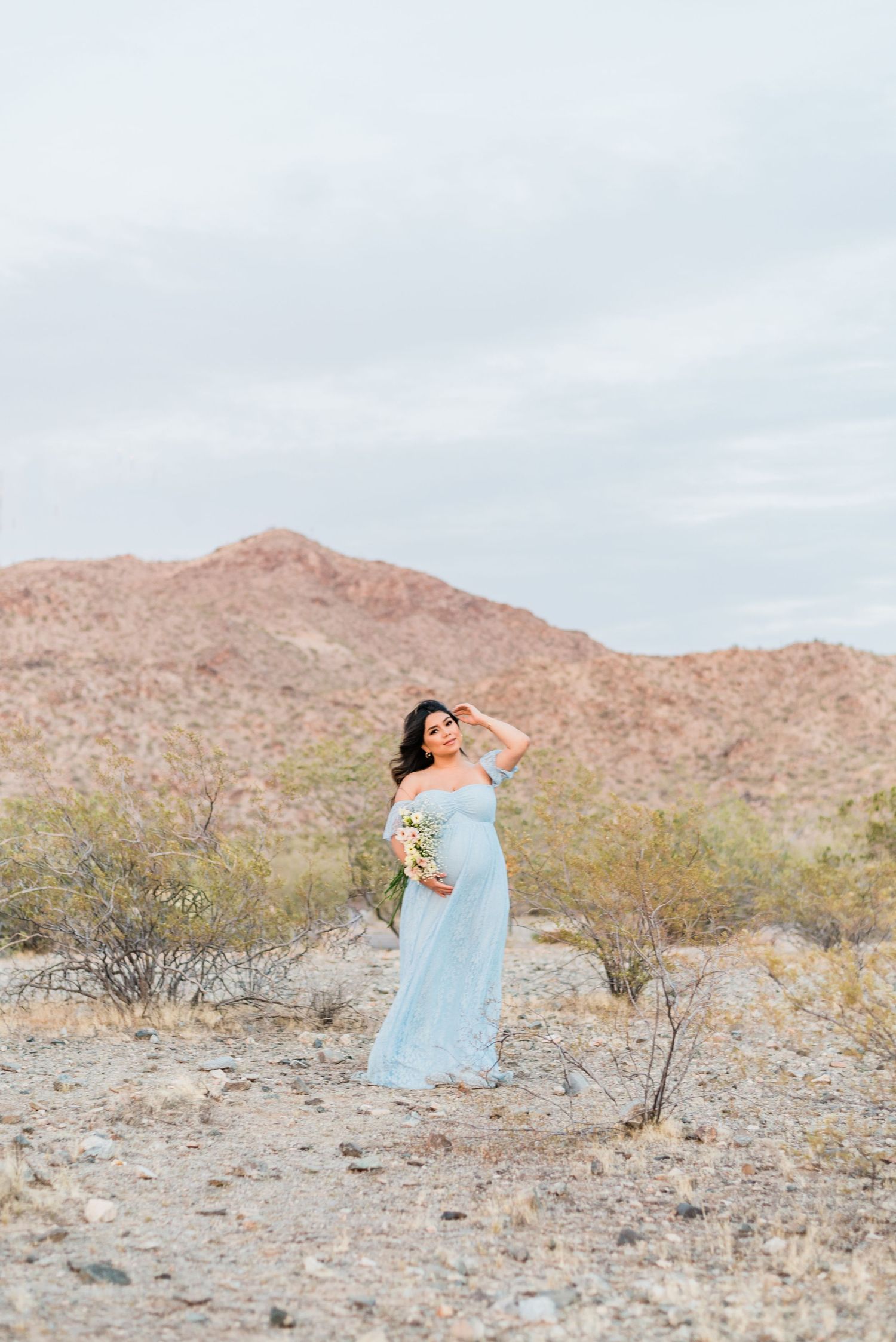 pregnant woman in a pale blue gown holds a bouquet among arizona desert mountains during a maternity session
