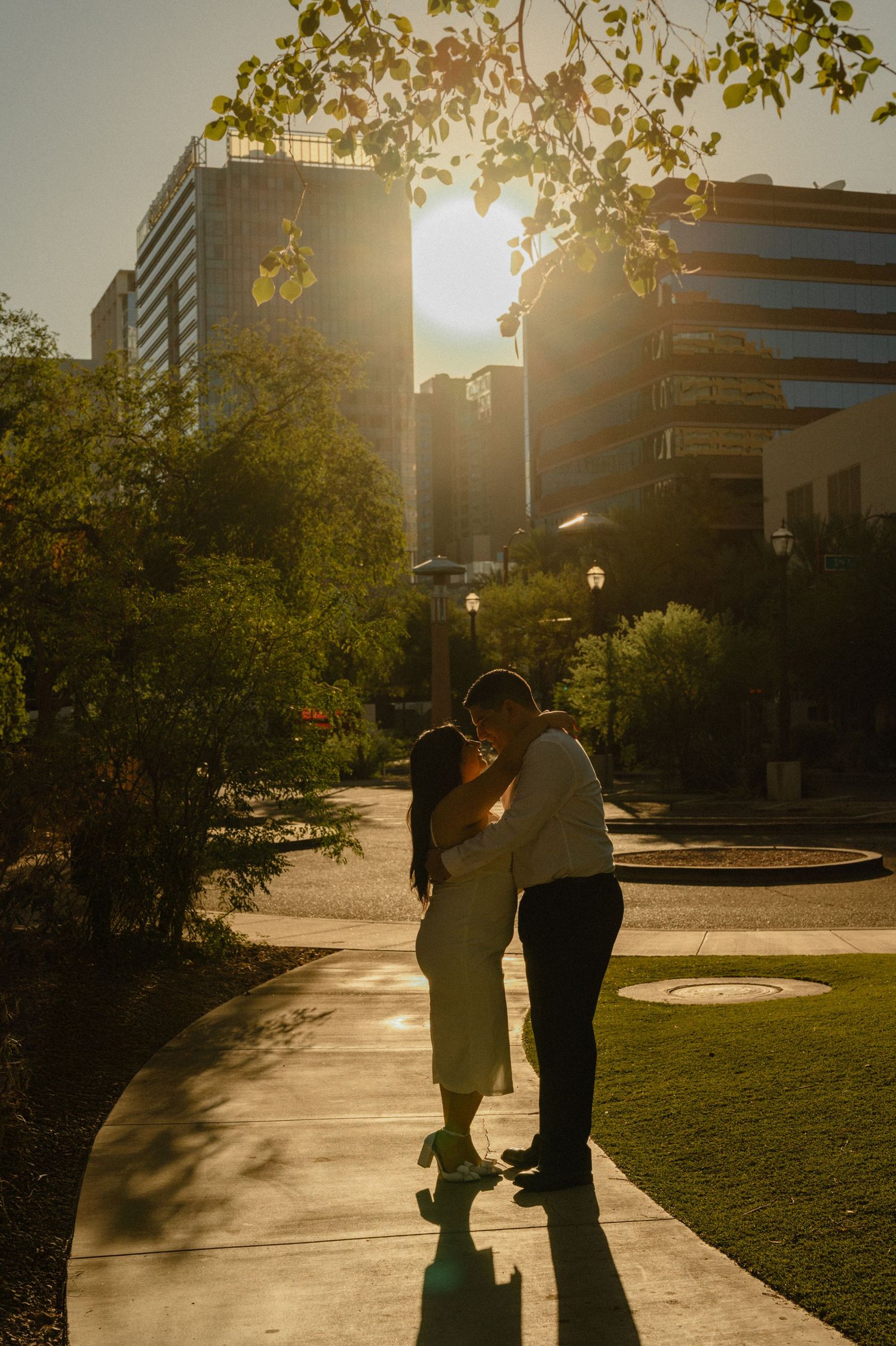 Couple sharing a romantic kiss at golden hour in an urban park with sunlight glowing between city buildings.