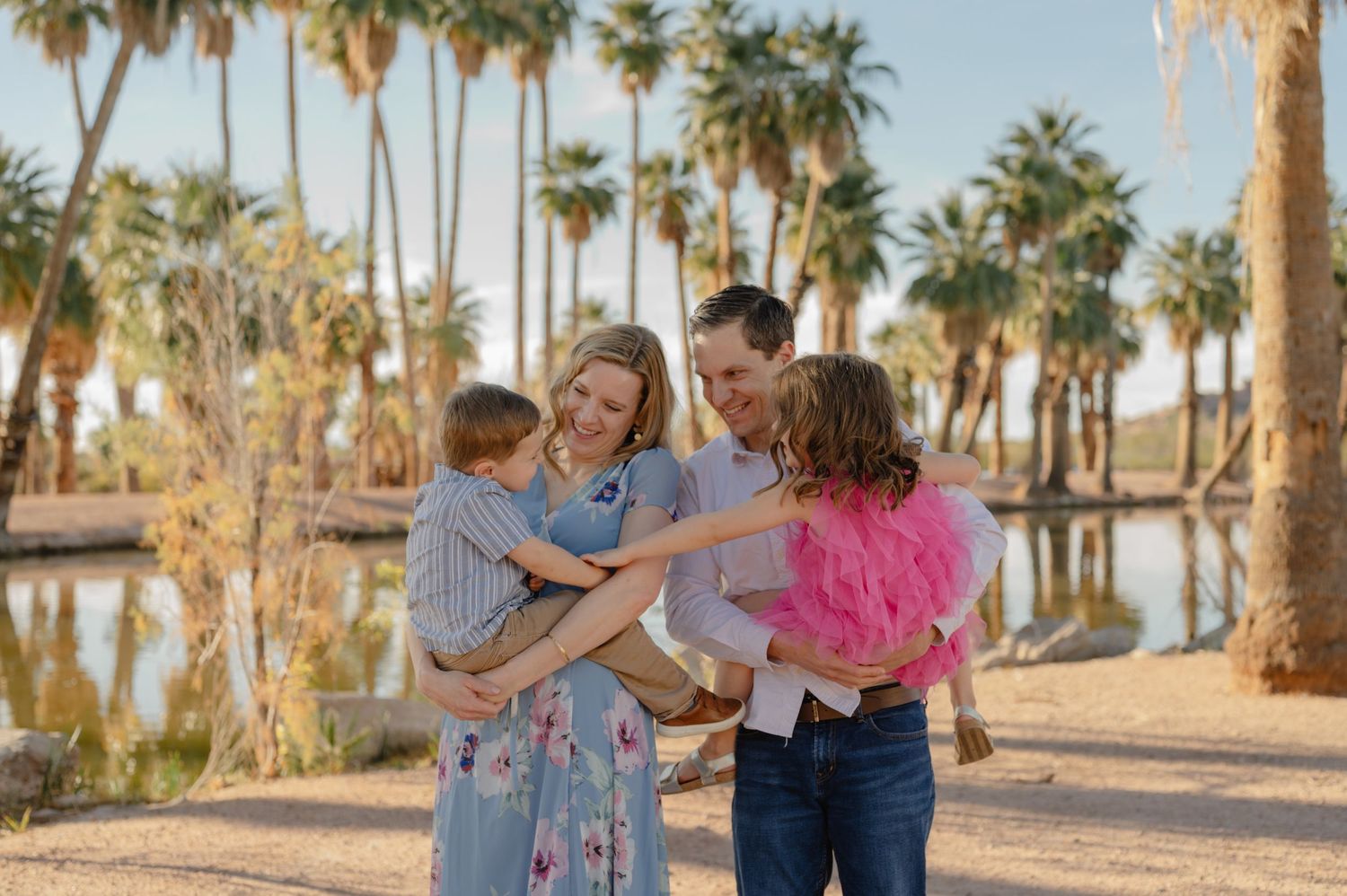 Family of four laughing together outdoors near a pond surrounded by tall palm trees, woman in blue dress, girl in pink tutu.