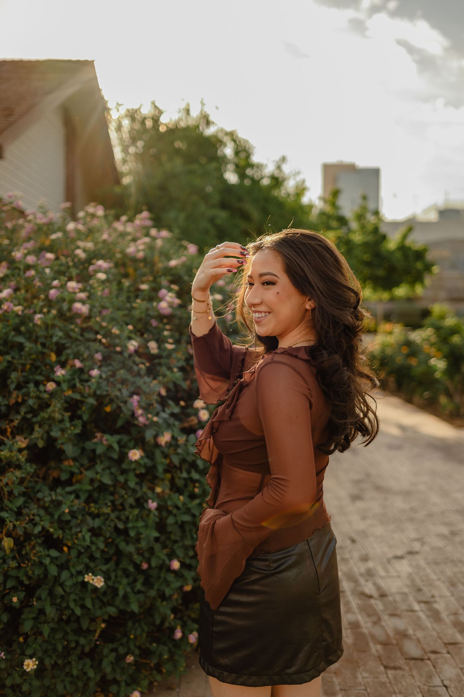 Young woman in brown top and leather skirt smiling outdoors near blooming pink flowers in golden hour light.