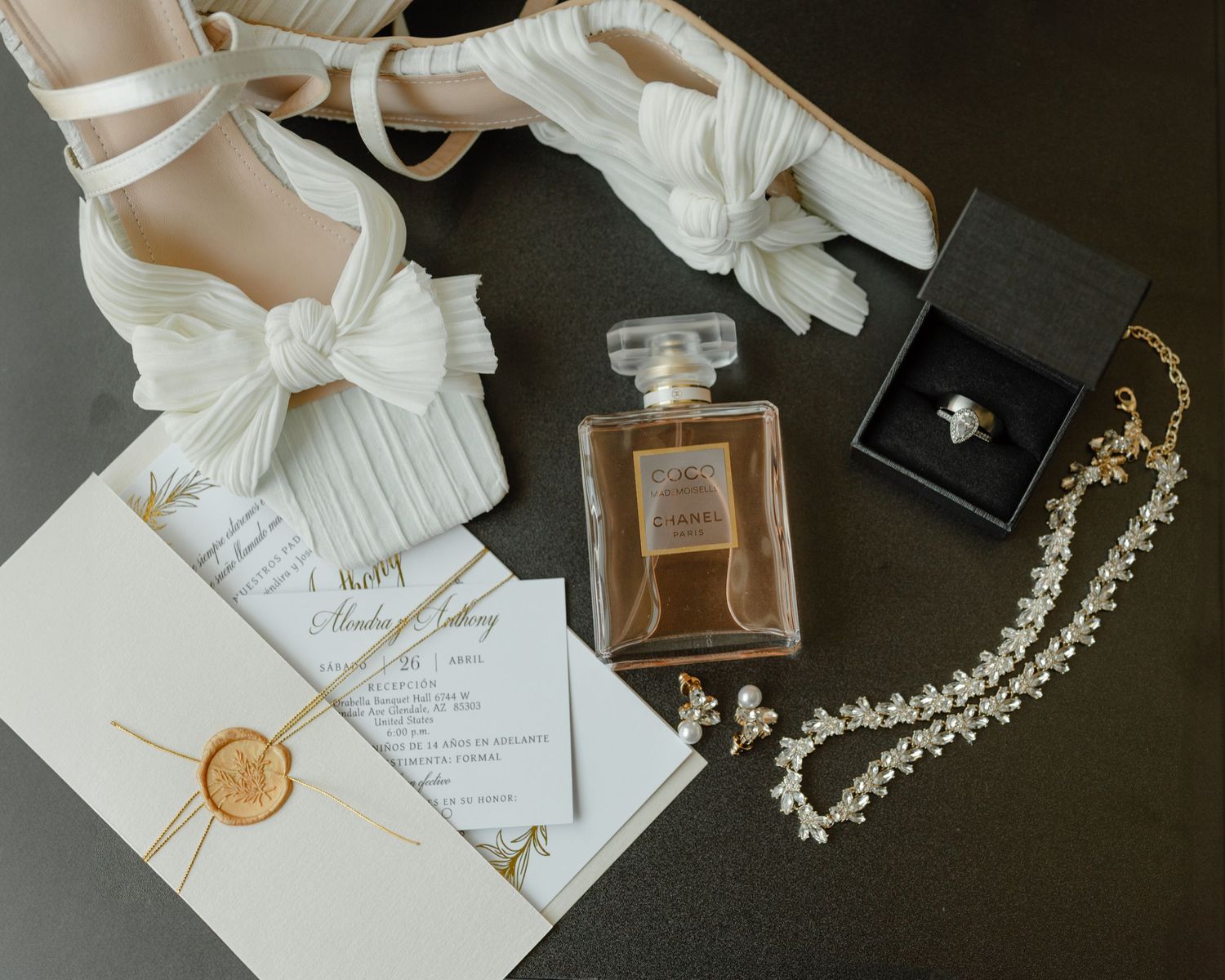 Flat lay of bridal accessories including heels, perfume, necklace, wedding invitation, and wax seal.