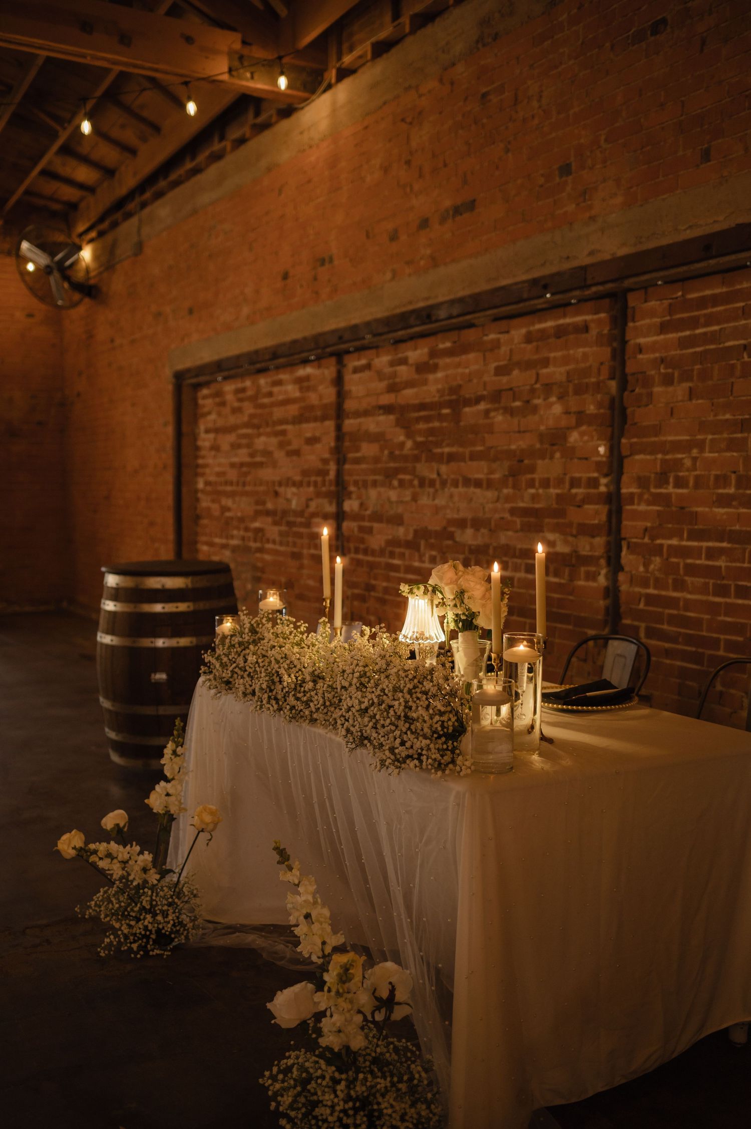 Romantic sweetheart table setup at wedding reception, with candles, white florals, and warm brick wall backdrop.
