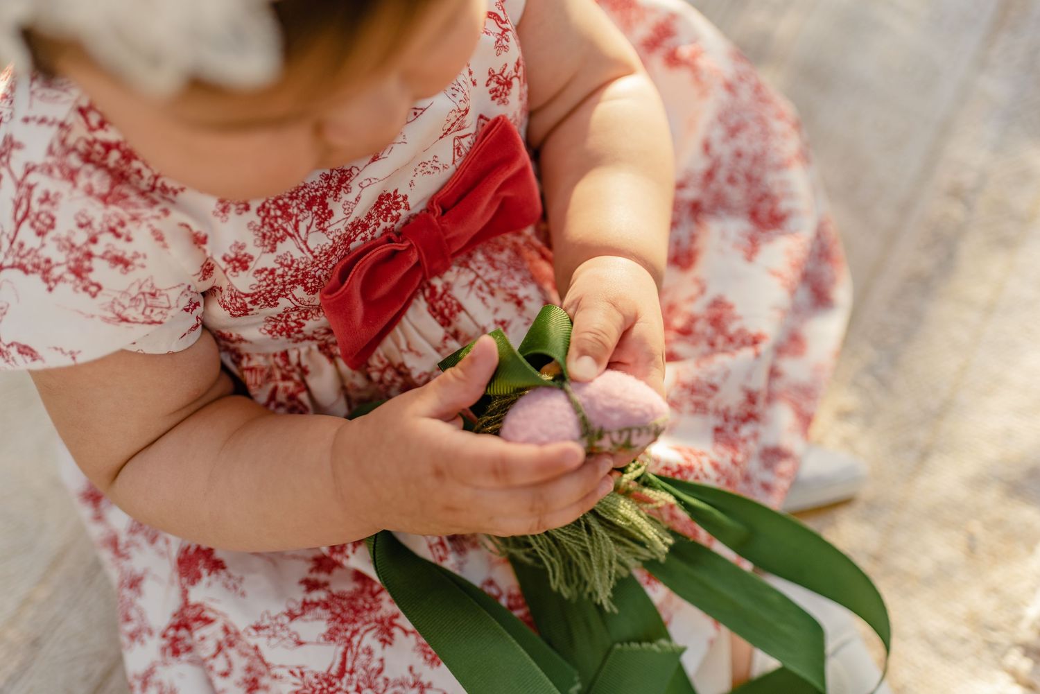 Baby in red floral dress holding a small pink flower bouquet with green leaves.
