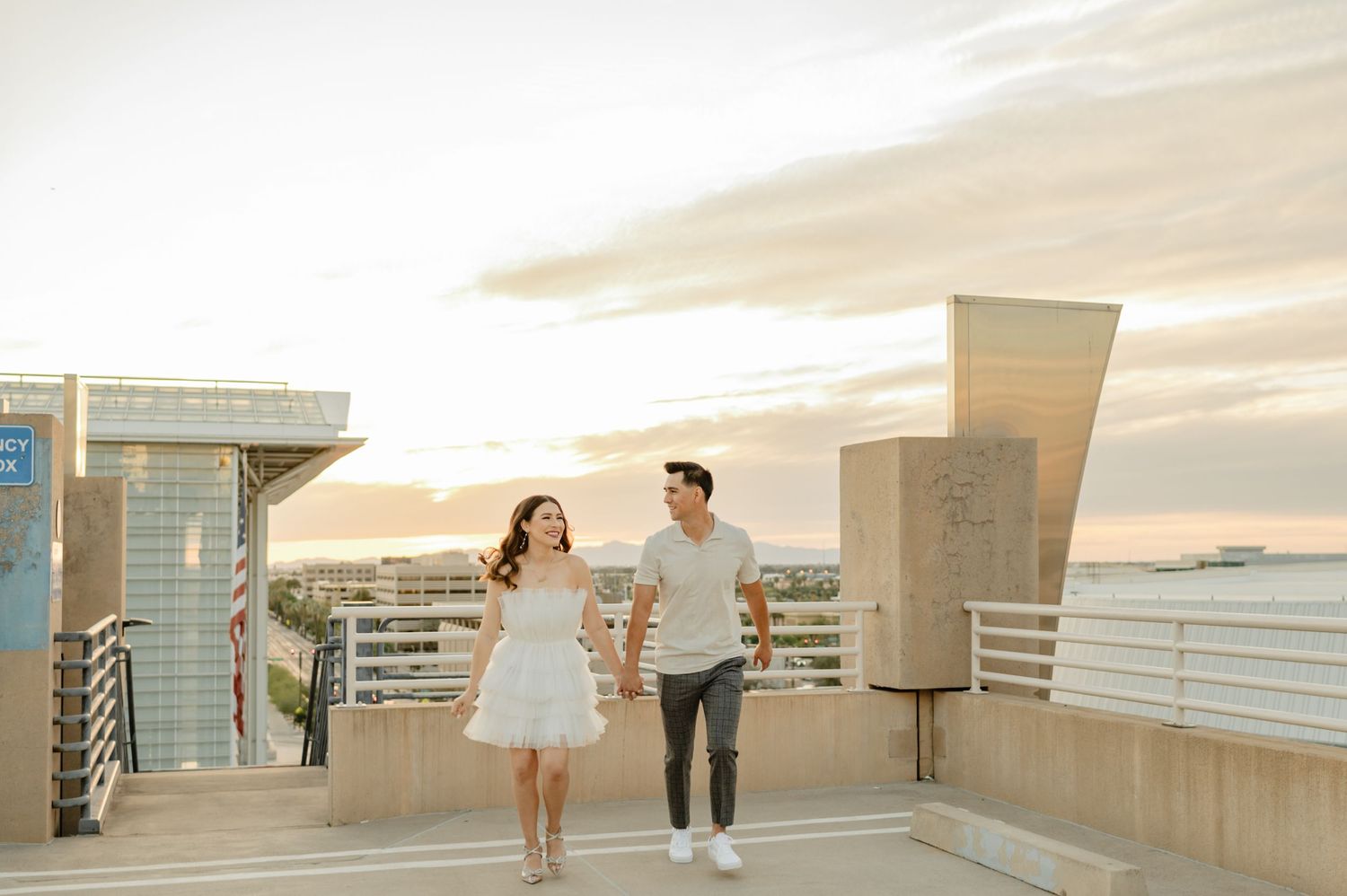 Couple holding hands walking on rooftop terrace at sunset with city skyline and golden sky in background.