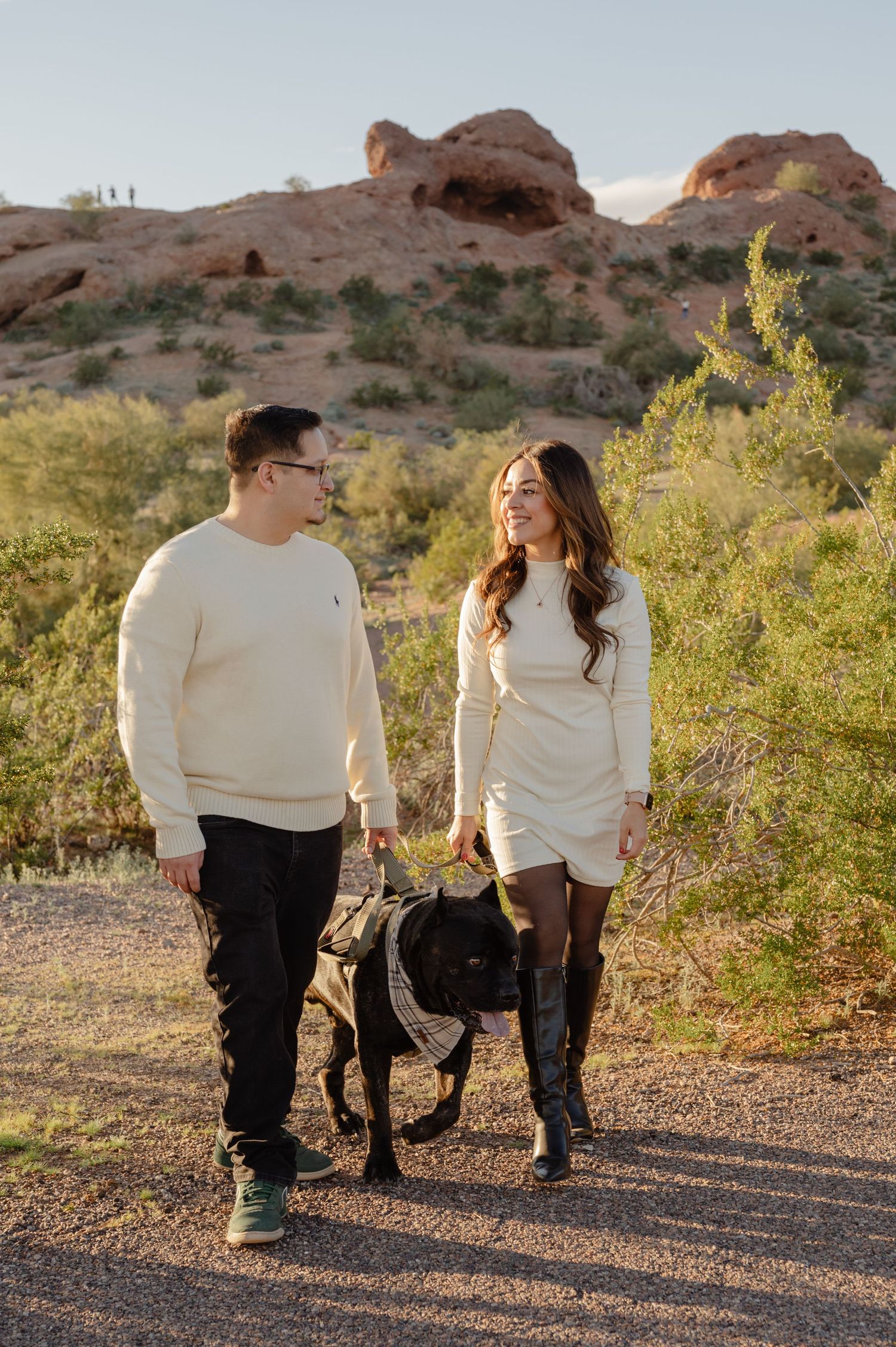 Couple walking a black dog on a desert trail with red rock formations in the background at golden hour.