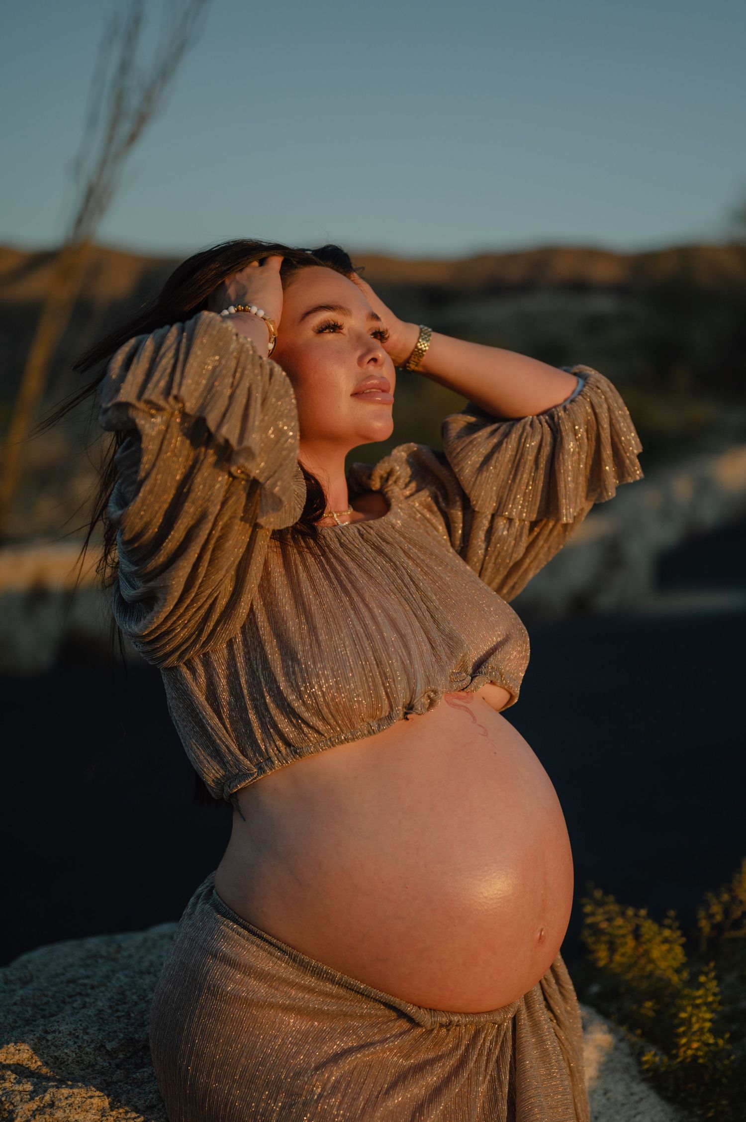 pregnant woman in a gold dress touches her hair at golden hour during a phoenix maternity photography session