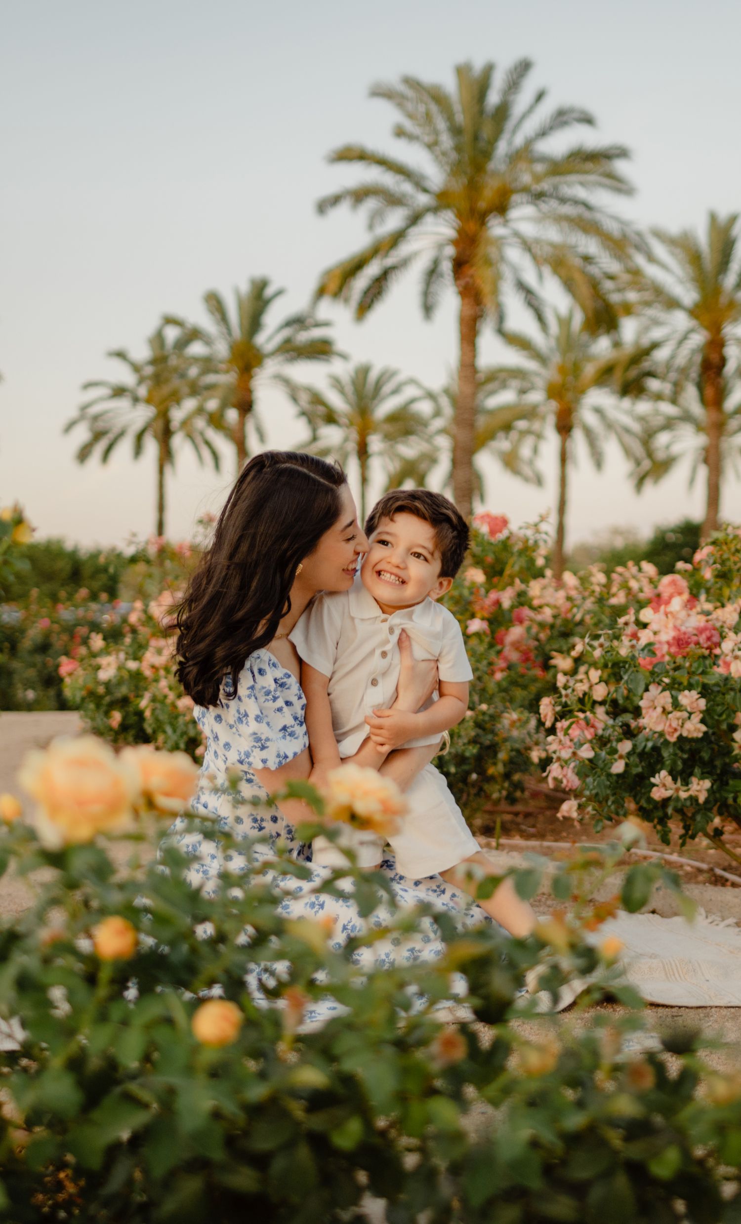 A mother kisses her smiling toddler among roses and palm trees during golden hour.