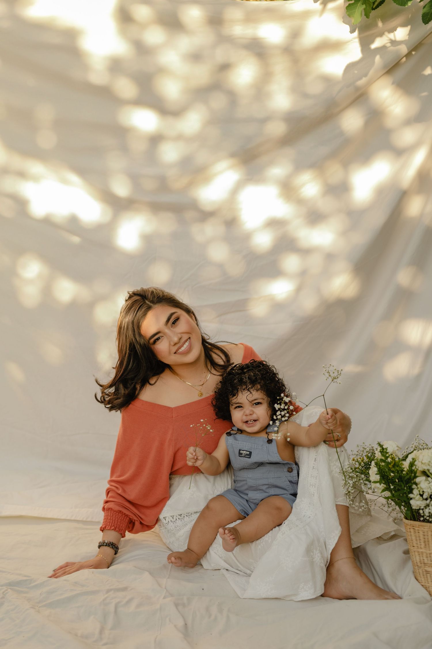 Smiling mother and curly-haired baby sitting against a sunlit white backdrop with flowers.