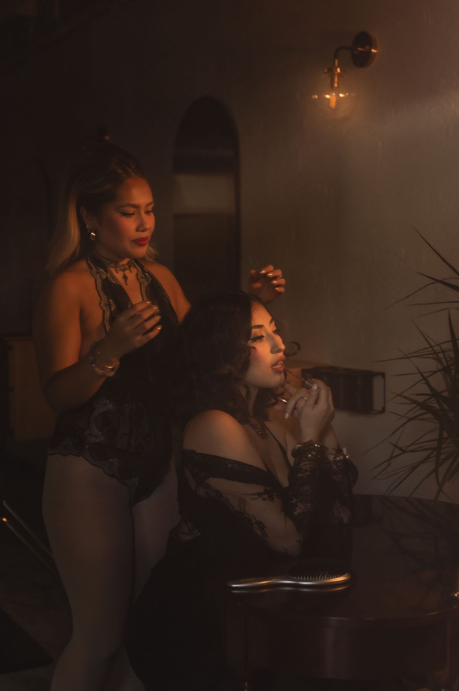 Two women sit in a dimly lit salon space, one styling the other's hair with a curling iron.