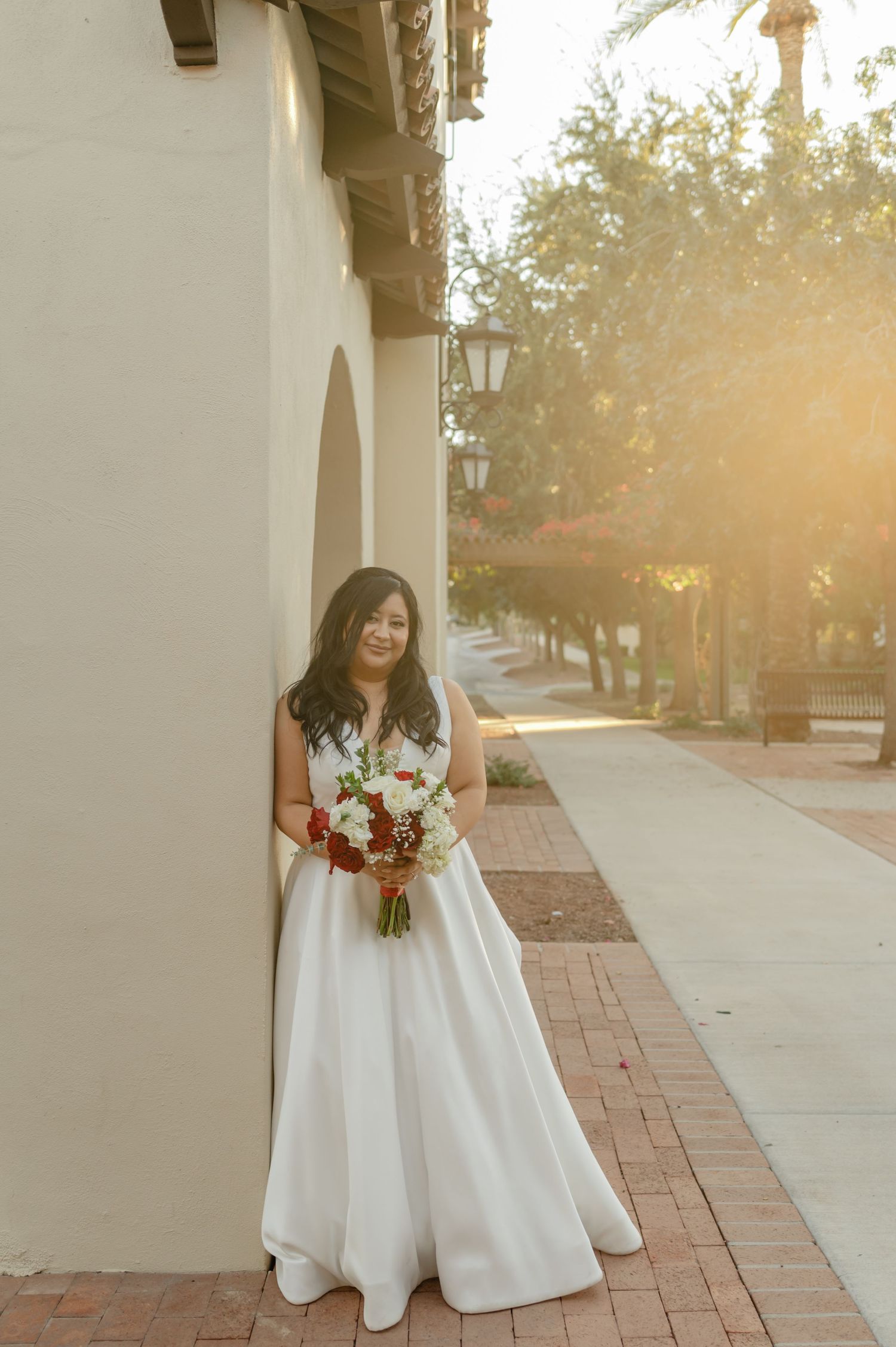 Bride stands in golden sunlight outside Spanish-style building holding bouquet.