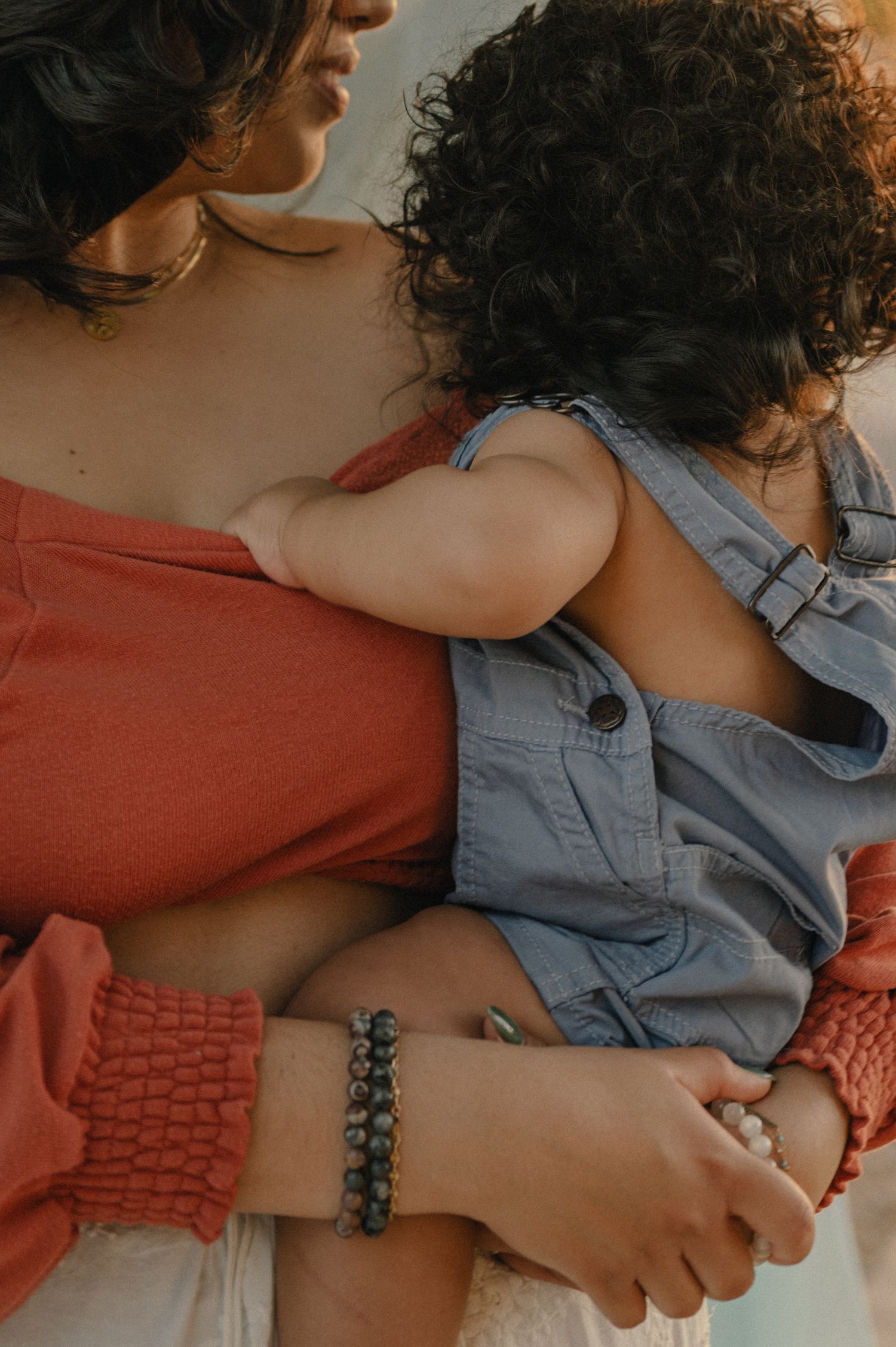 Close-up of a mother in red top holding a young child in denim overalls, wearing beaded bracelets.