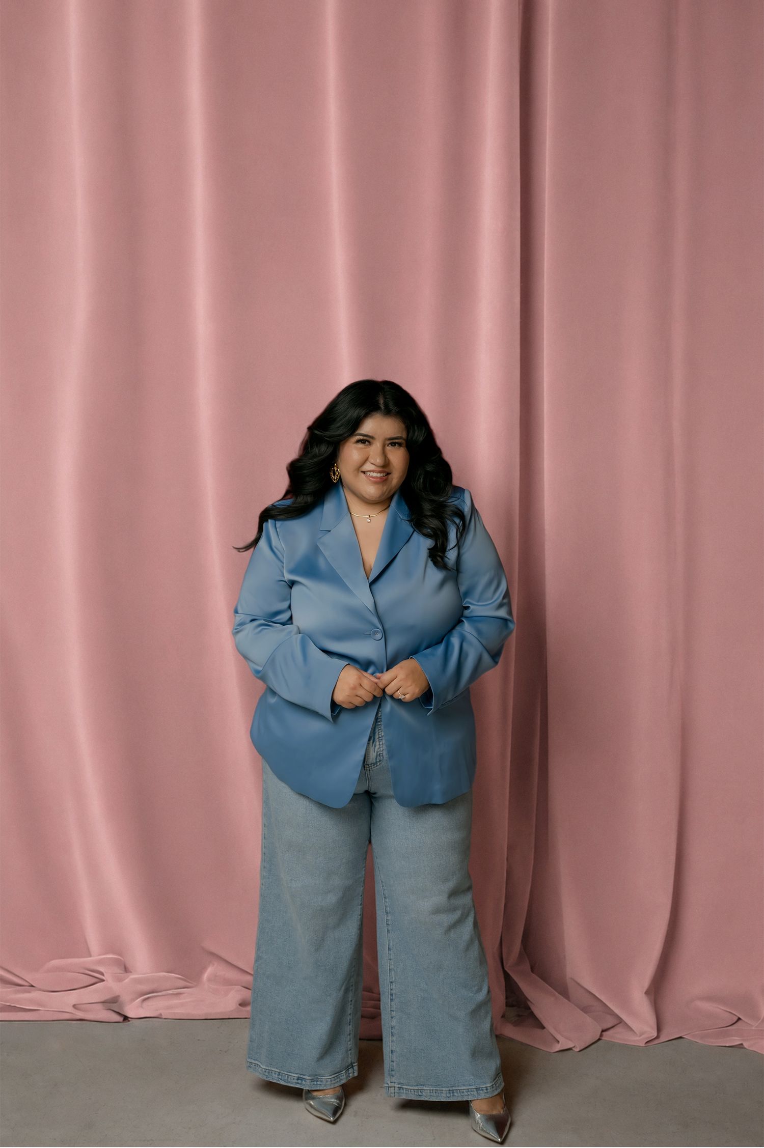Sharom Rosas, founder of Shamari Visuals, poses in a blue blazer and wide leg jeans against a pink drape backdrop.