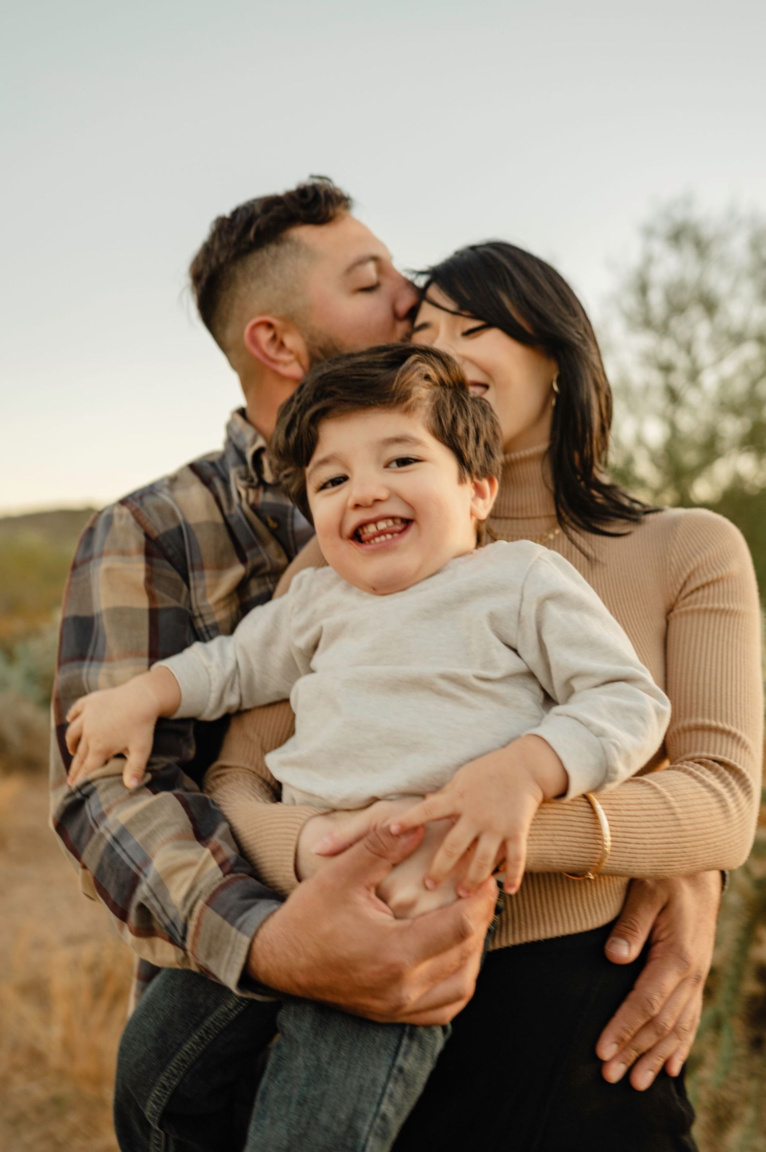 Happy parents kissing and holding their laughing toddler outdoors in a desert setting.