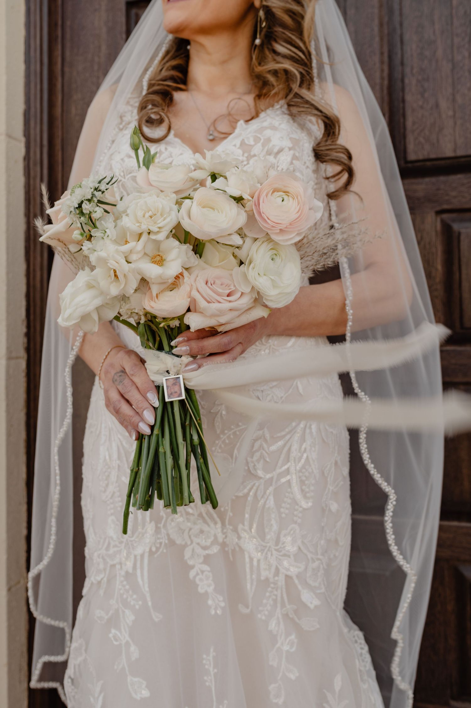 Bride holding blush pink and white bouquet in lace wedding dress and veil, framed by dark wood doors.