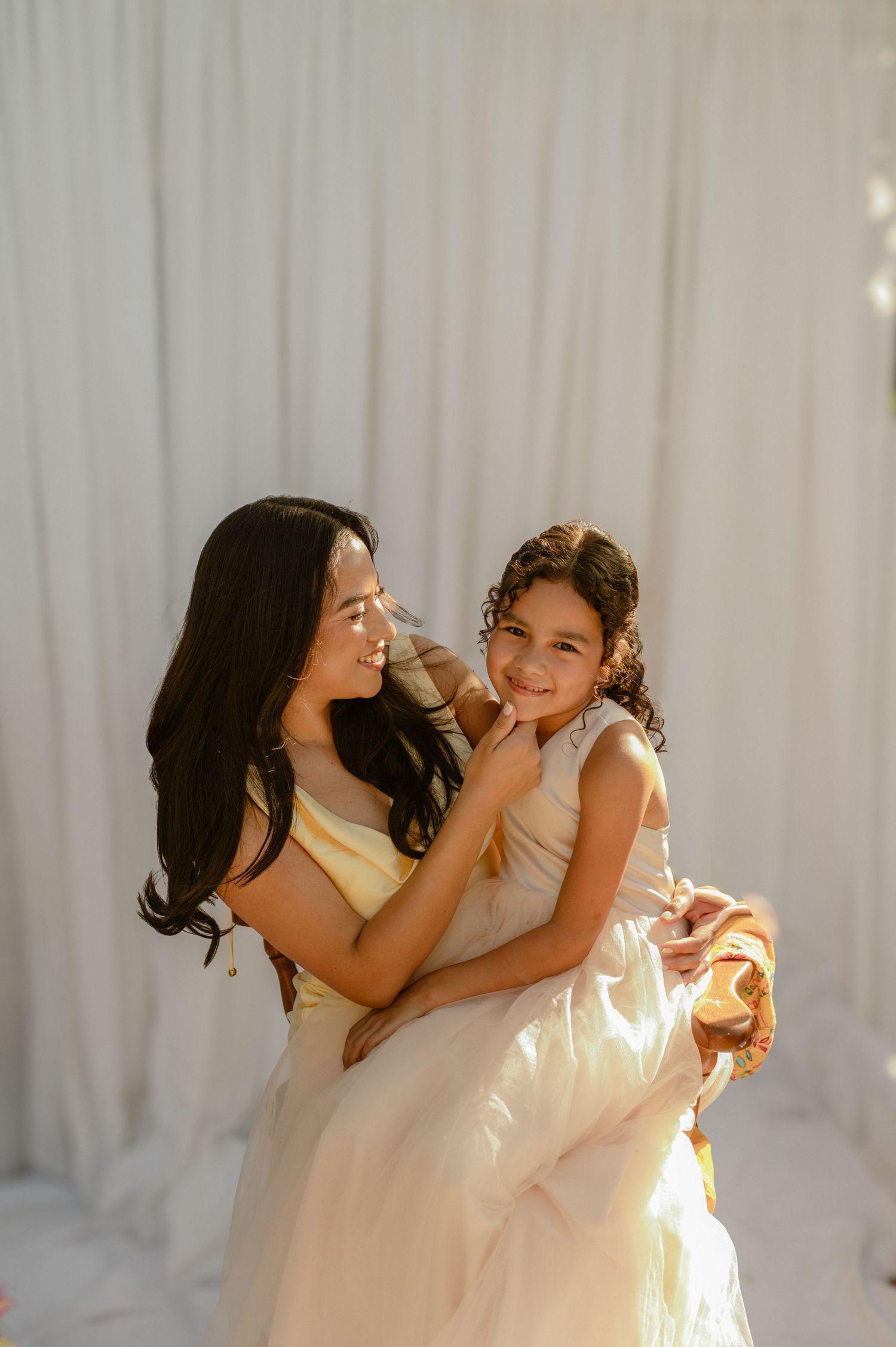 Mommy and me session in Phoenix, AZ, mother and daughter smiling together in soft flowing dresses against a draped backdrop.