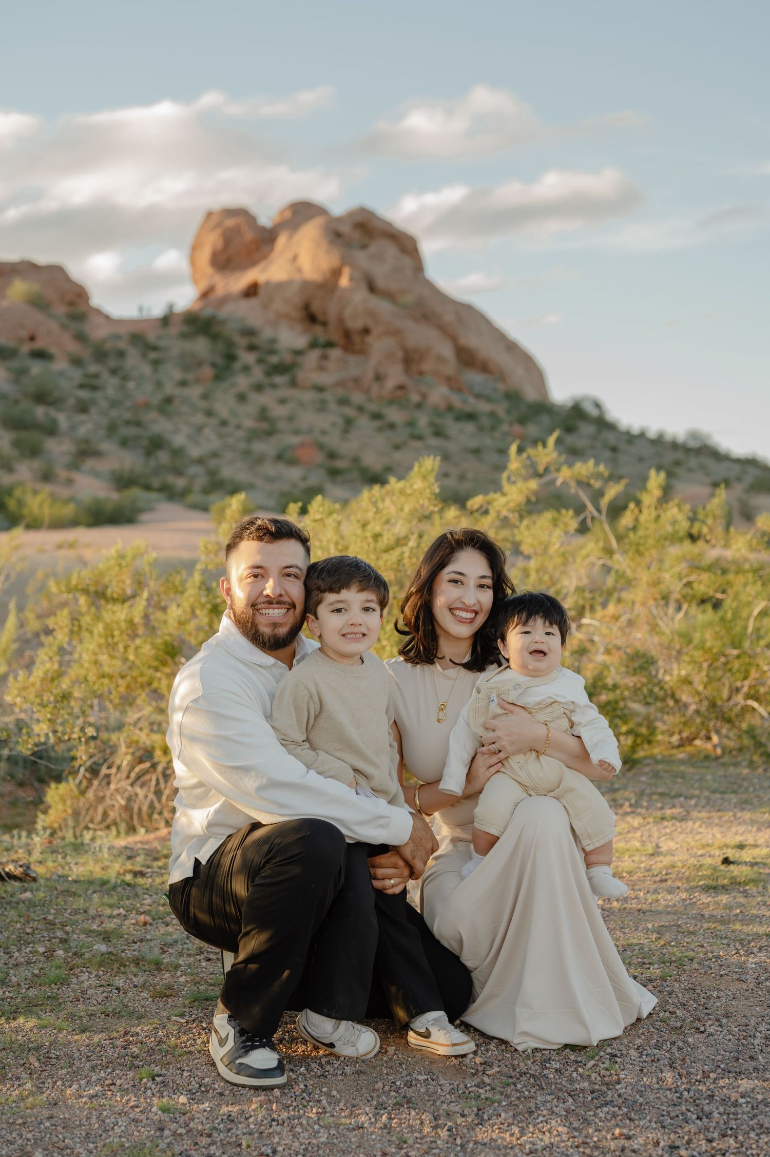 Family of four poses smiling outdoors near red rock formations in desert landscape during golden hour.