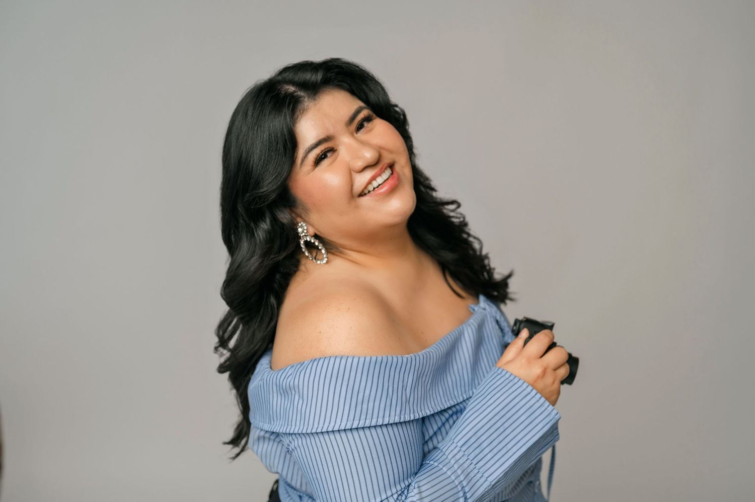 Smiling woman with long dark hair wearing a light blue off-the-shoulder top against a gray background.