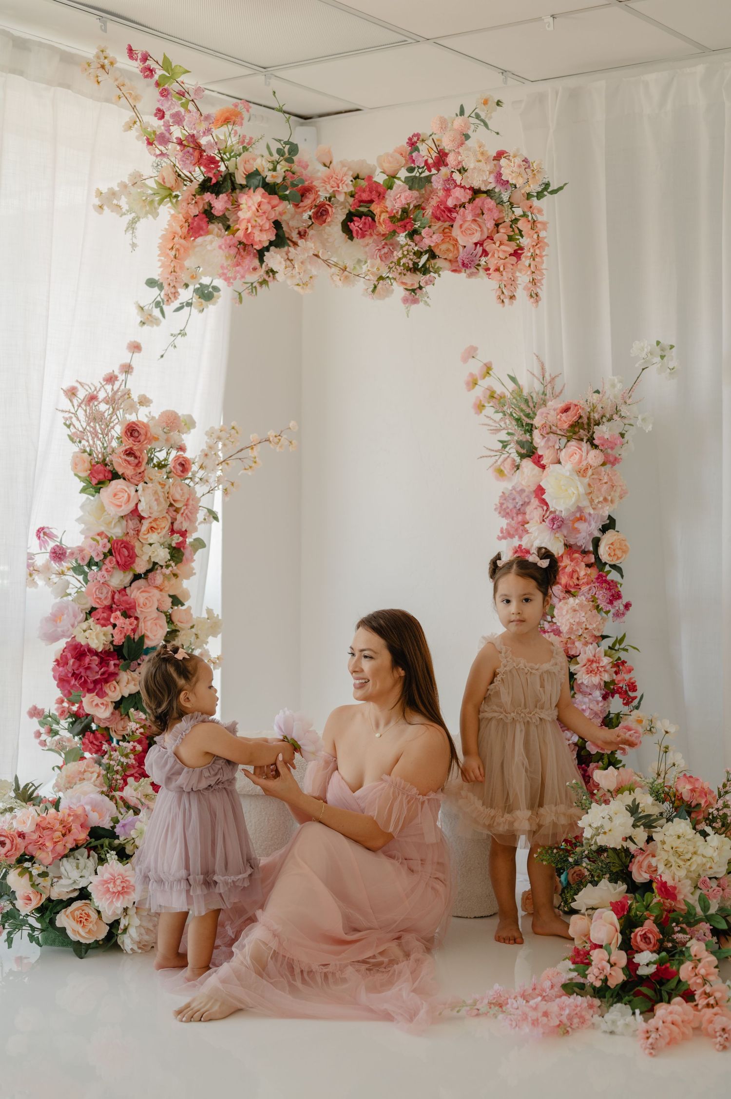 Mother in pink gown sitting under floral arch with two young daughters in studio.