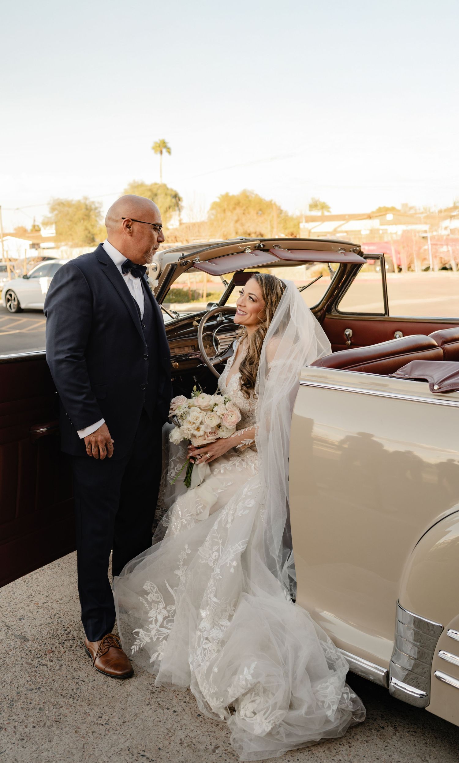 Groom helping bride out of vintage cream convertible during golden hour, romantic and classic wedding moment.