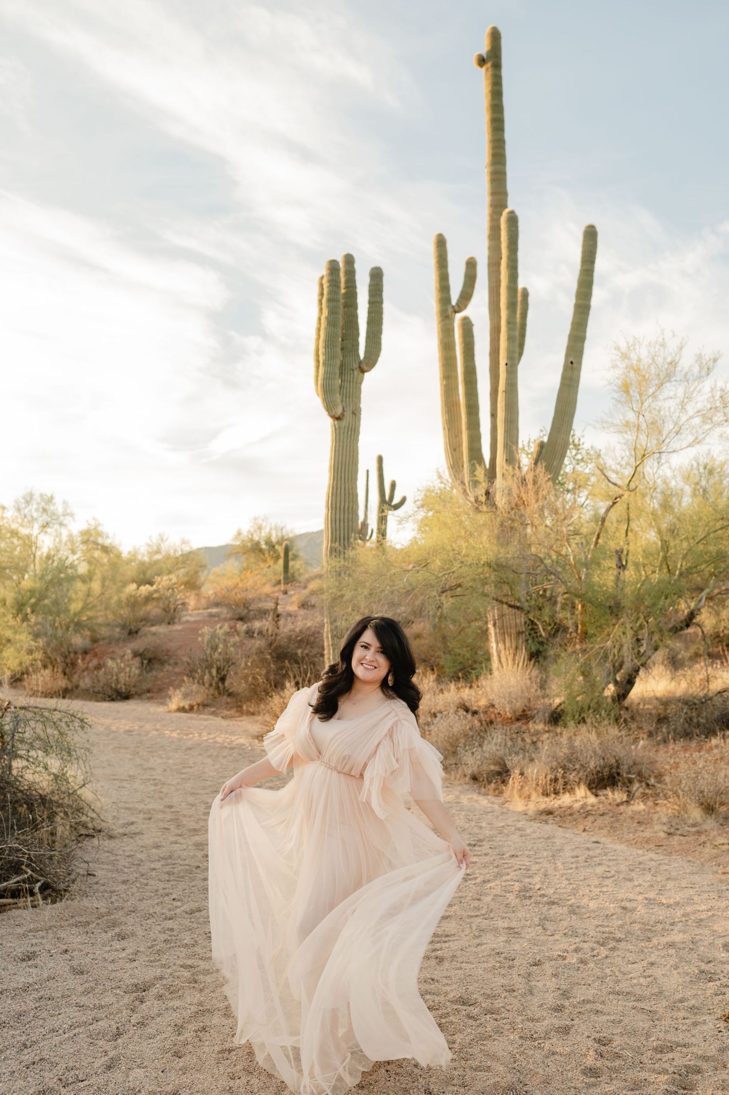 Pregnant woman in flowing blush tulle gown twirling in Arizona desert with saguaro cacti.