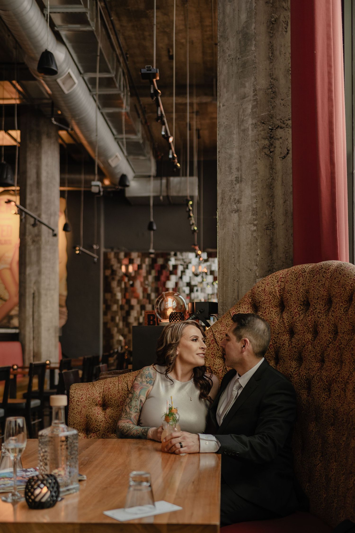Couple in wedding attire sharing an intimate moment in a moody industrial restaurant booth with Edison bulb lighting.