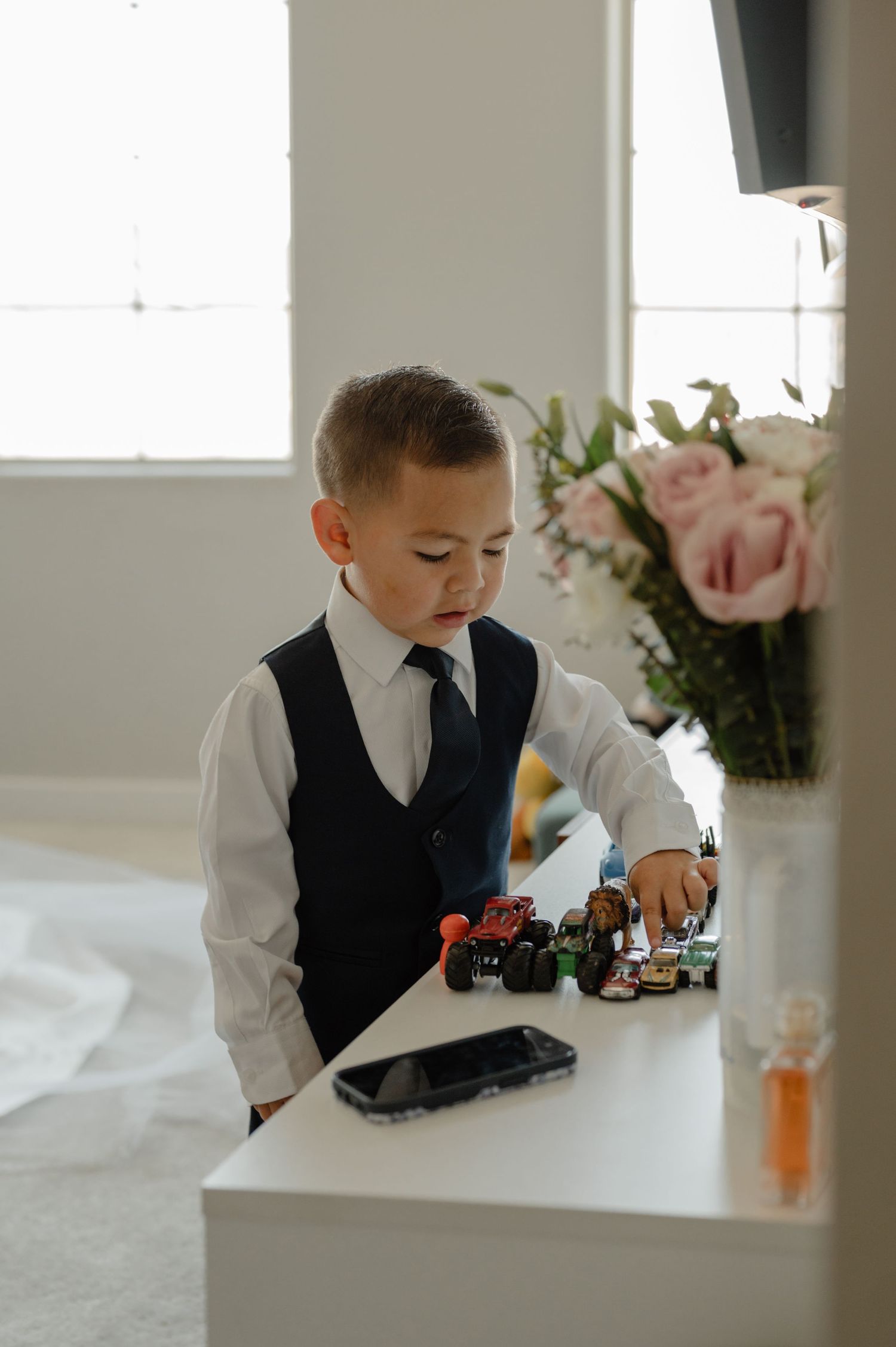 A young boy in a formal vest and tie plays with toy trucks on a white dresser during an indoor session.